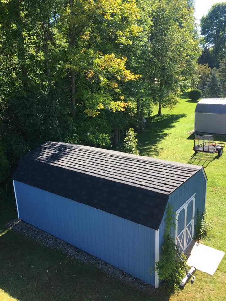 Blue barn with black roof in a grassy yard, surrounded by trees and a small shed nearby