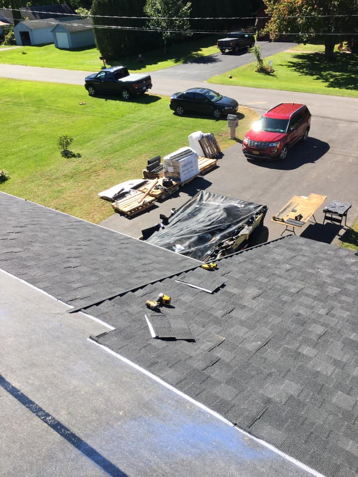 Roofing materials and workers on a house roof, with cars and equipment in the driveway below