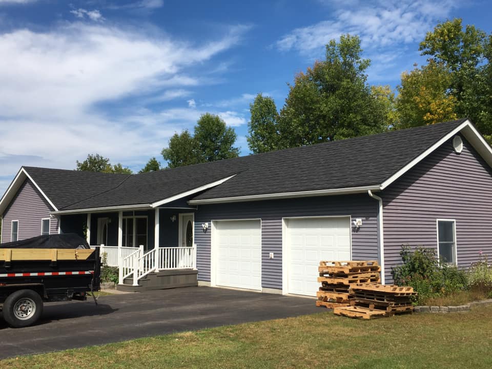 Single-story gray house with white garage doors, a driveway, and stacked lumber under a blue sky