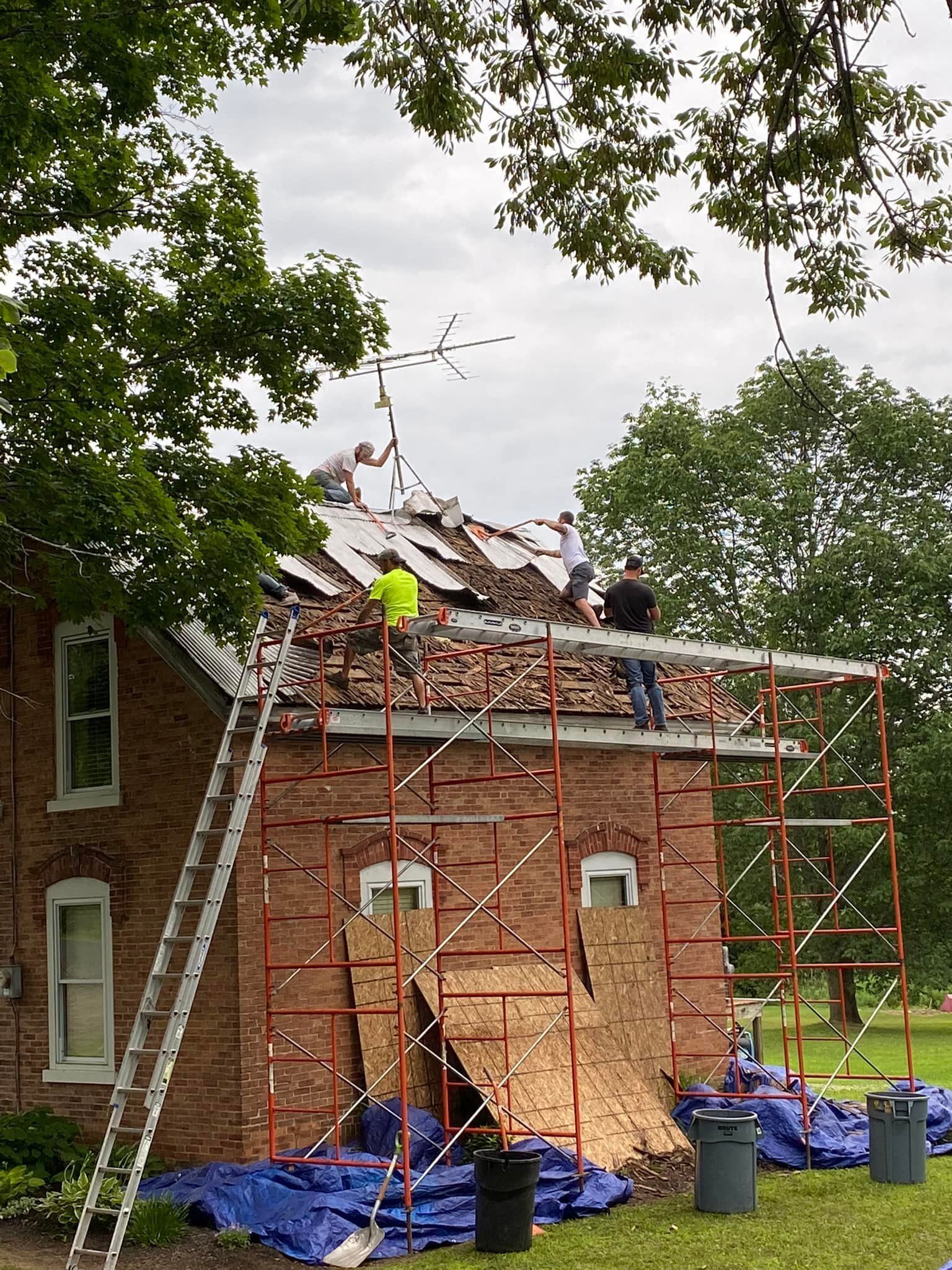 Workers repairing a brick roof with scaffolding and blue tarps under trees