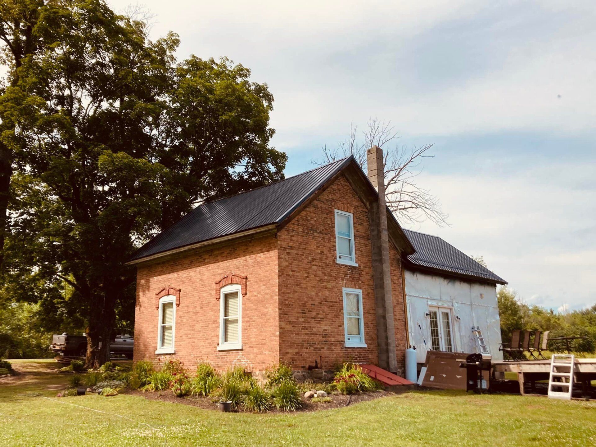 Red brick cottage with white side wall, dark roof, and large trees in a sunny rural yard