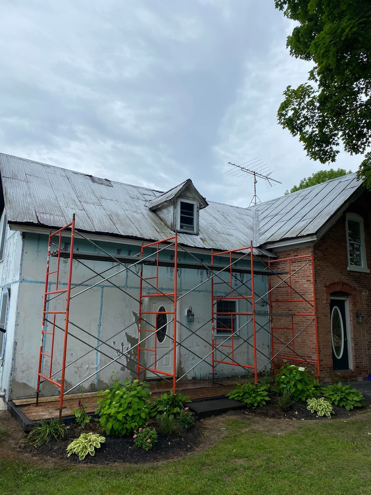 House exterior under renovation with red scaffolding, gray roof, and brick and white siding.