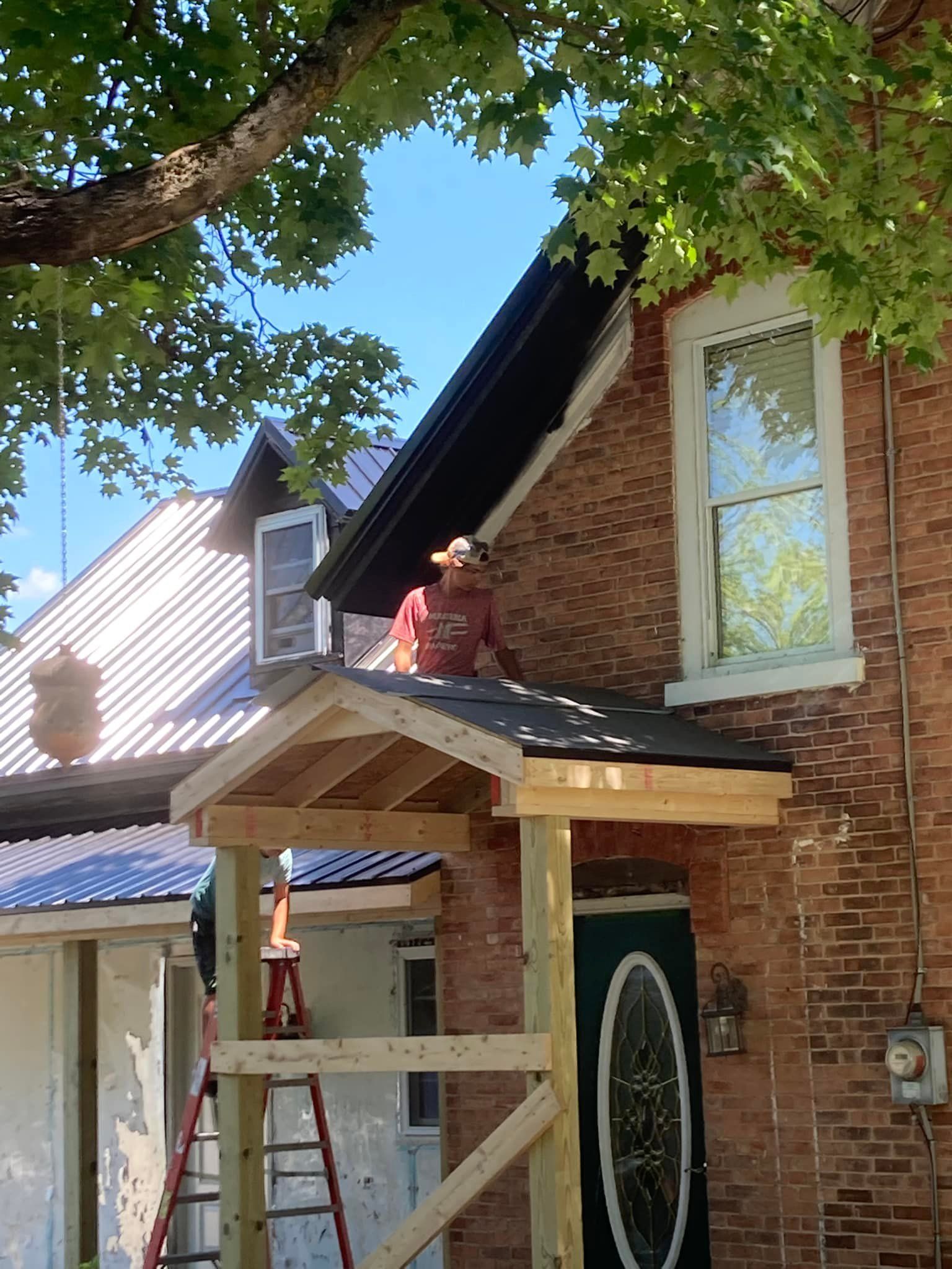 Person working on a roof over a front porch beside a brick house with a ladder and tree shade