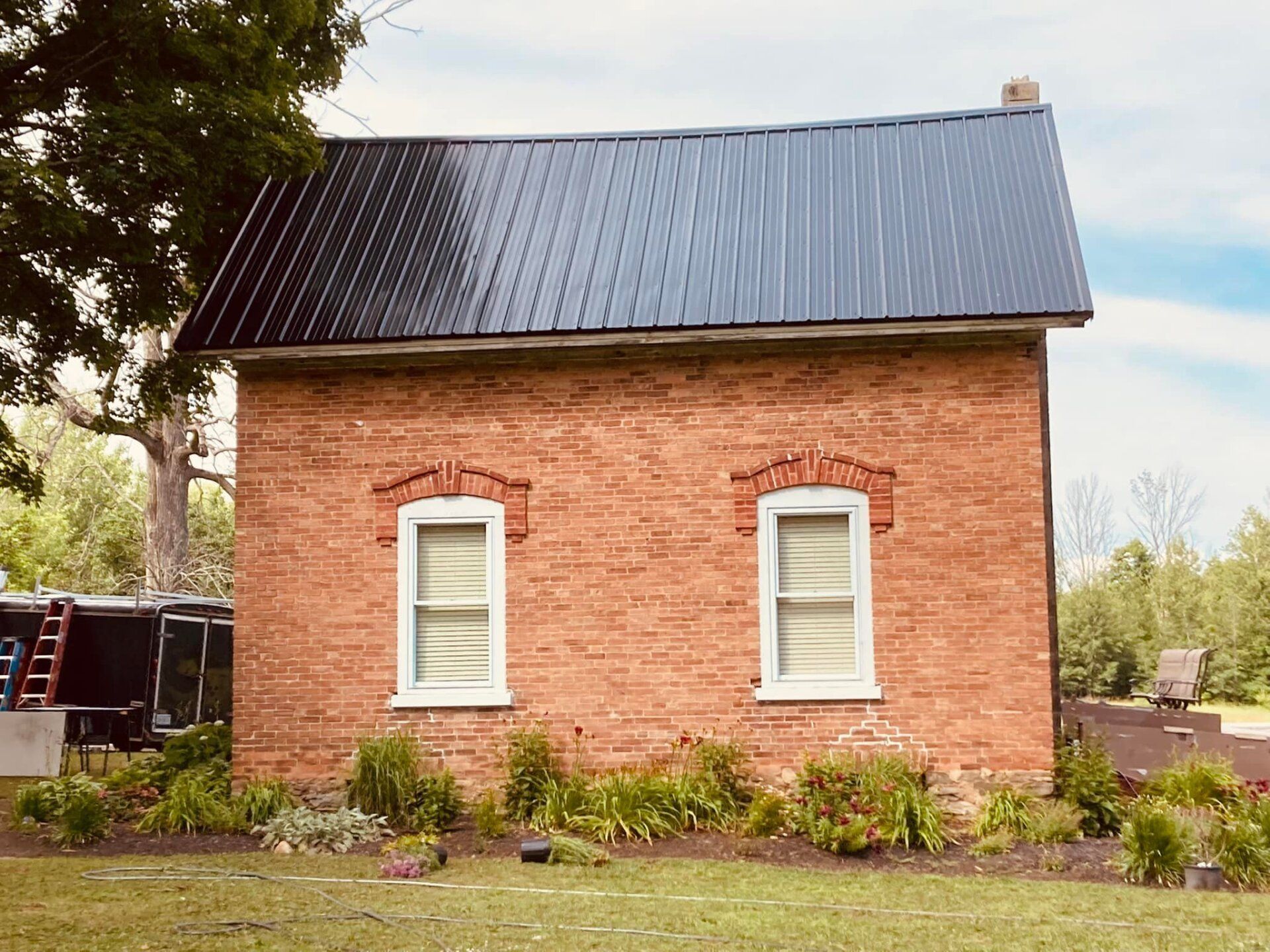 Small red brick house with a dark metal roof, white windows, and garden plants beside it.