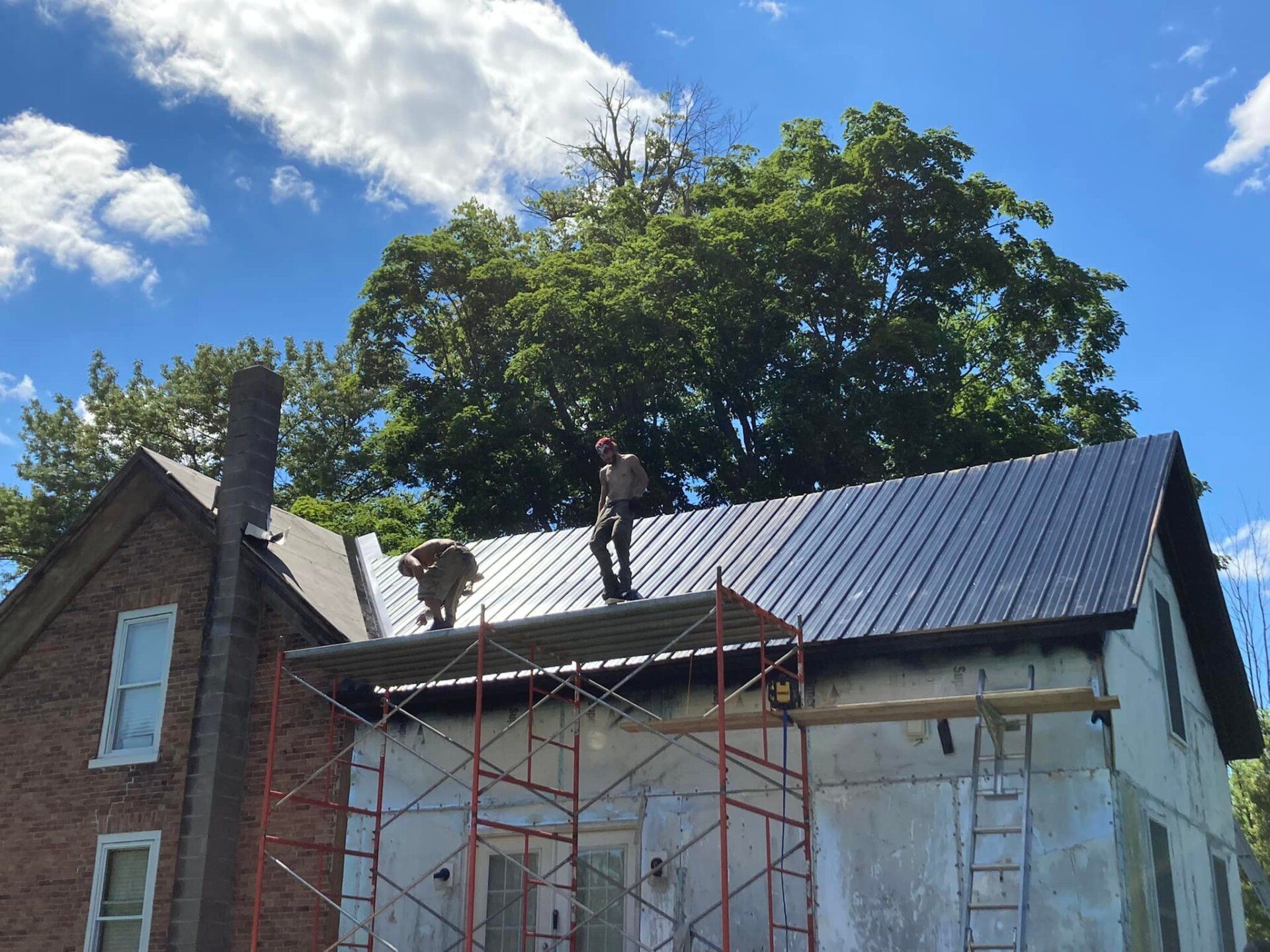 Workers on scaffolding installing a dark metal roof on a brick house under a blue sky