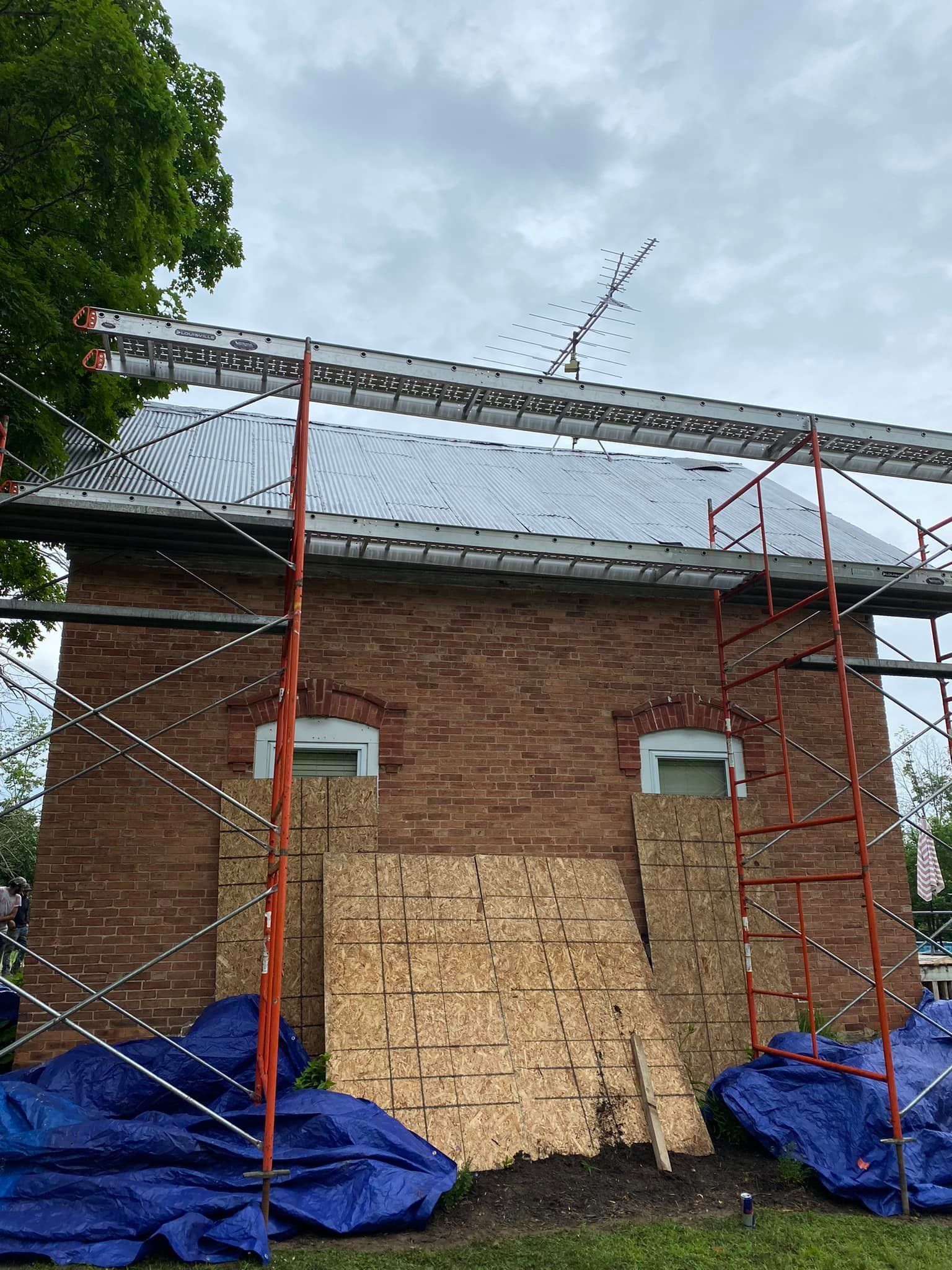Brick house under construction with scaffolding, boarded windows, and blue tarps at the base