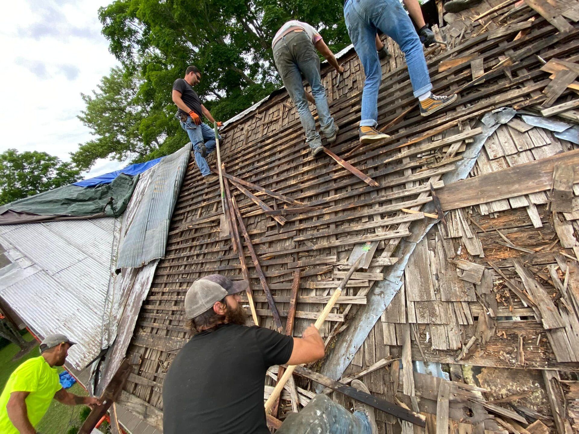 Workers repairing a damaged roof on a house, with shingles removed and ladders in place.