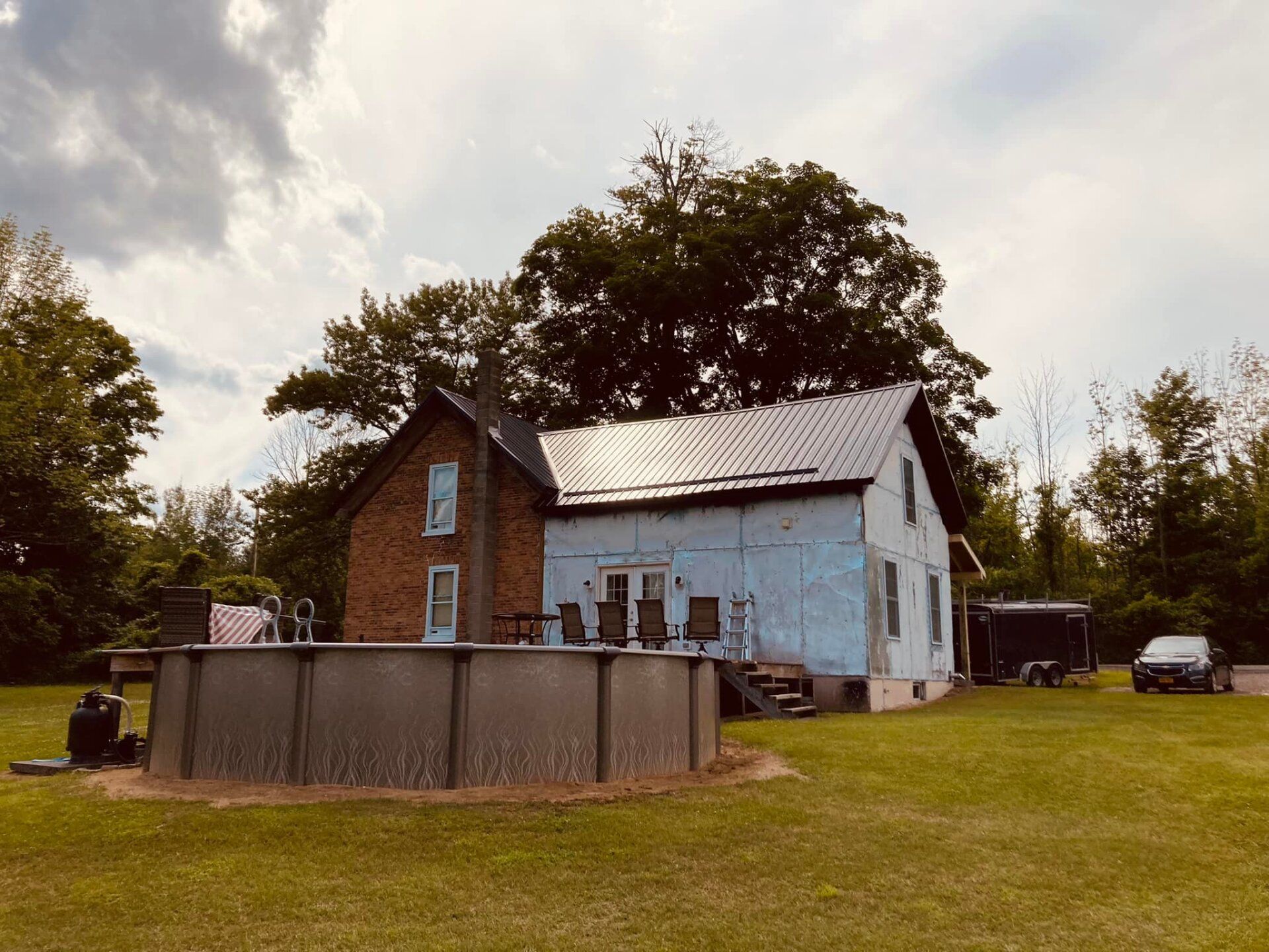 Old farmhouse with white siding and brick chimney beside a round above-ground pool on a grassy lawn.