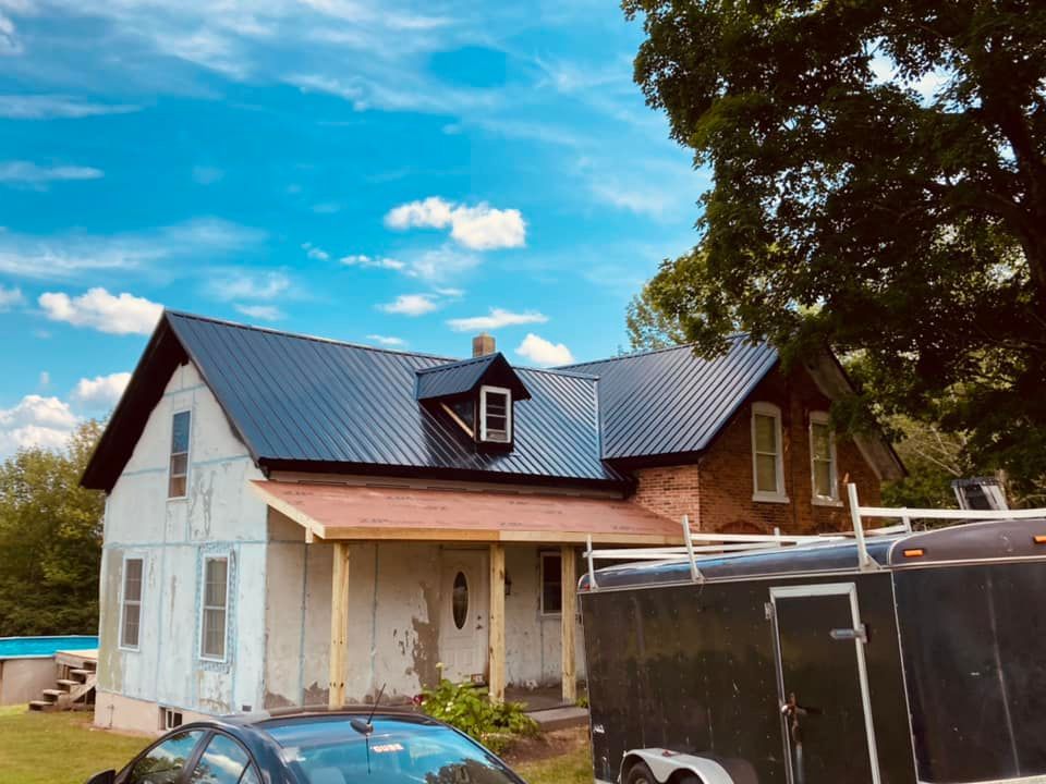 Partially renovated house with blue metal roof, porch, and trailer in a rural yard