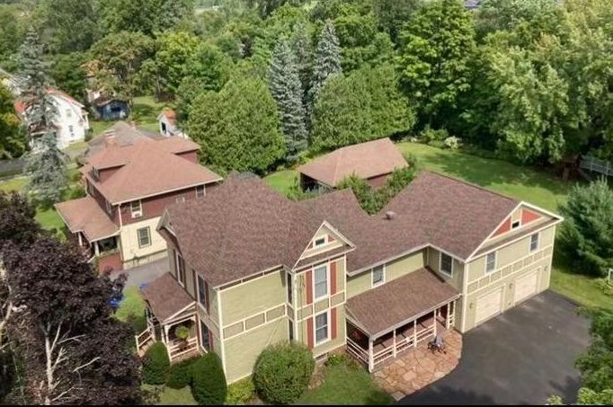 Aerial view of a large beige house with brown roof, surrounded by trees and a driveway.