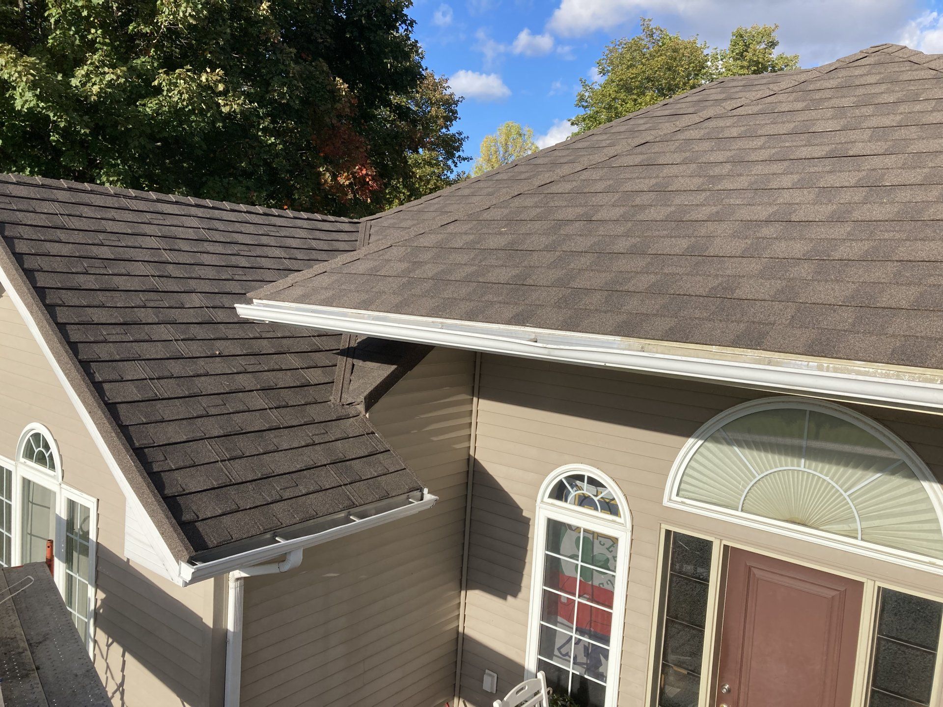 Suburban house roof with dark shingles and tan siding under a clear blue sky