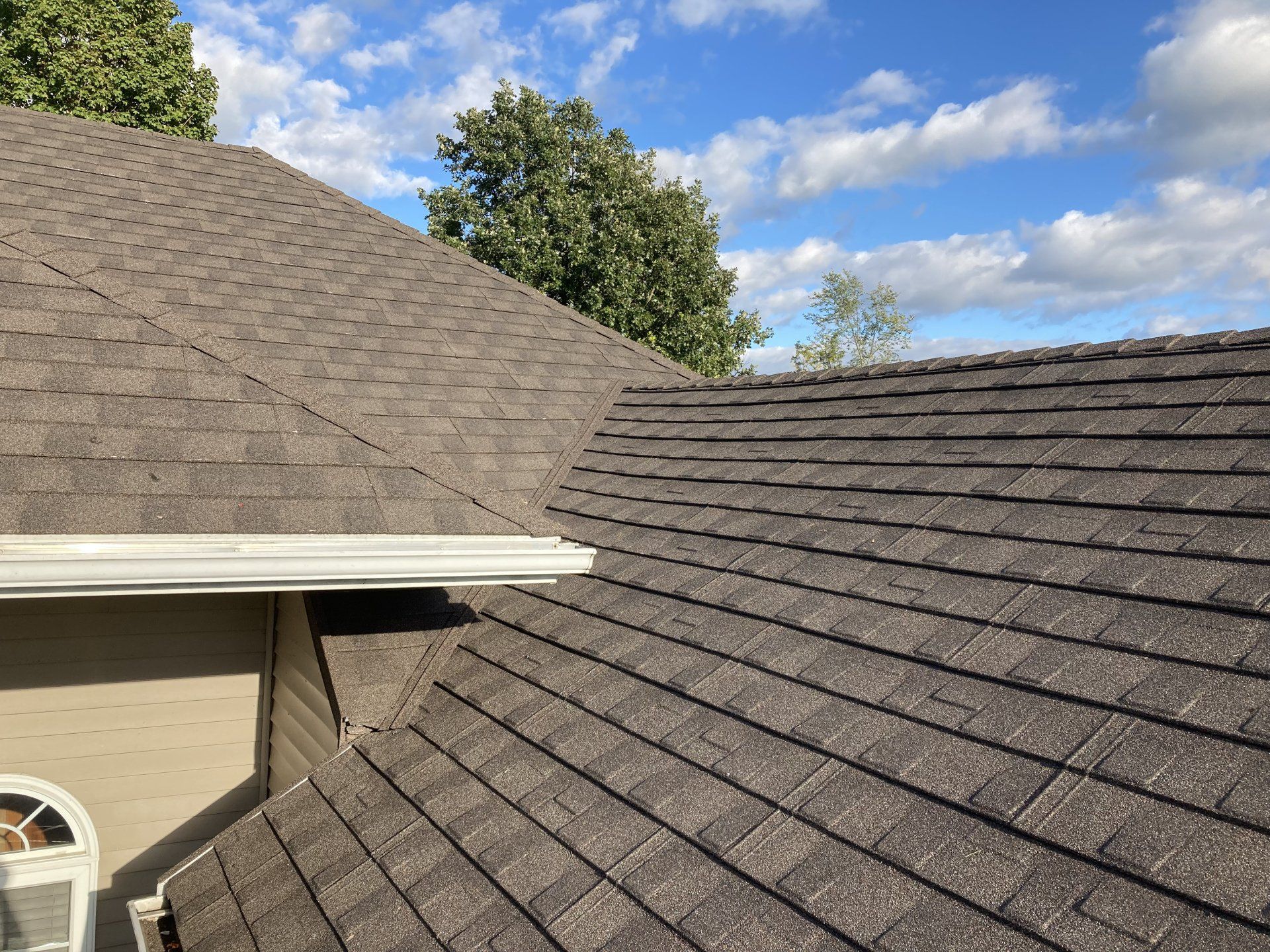 Residential roof with gray shingles and a white gutter under a blue sky with clouds