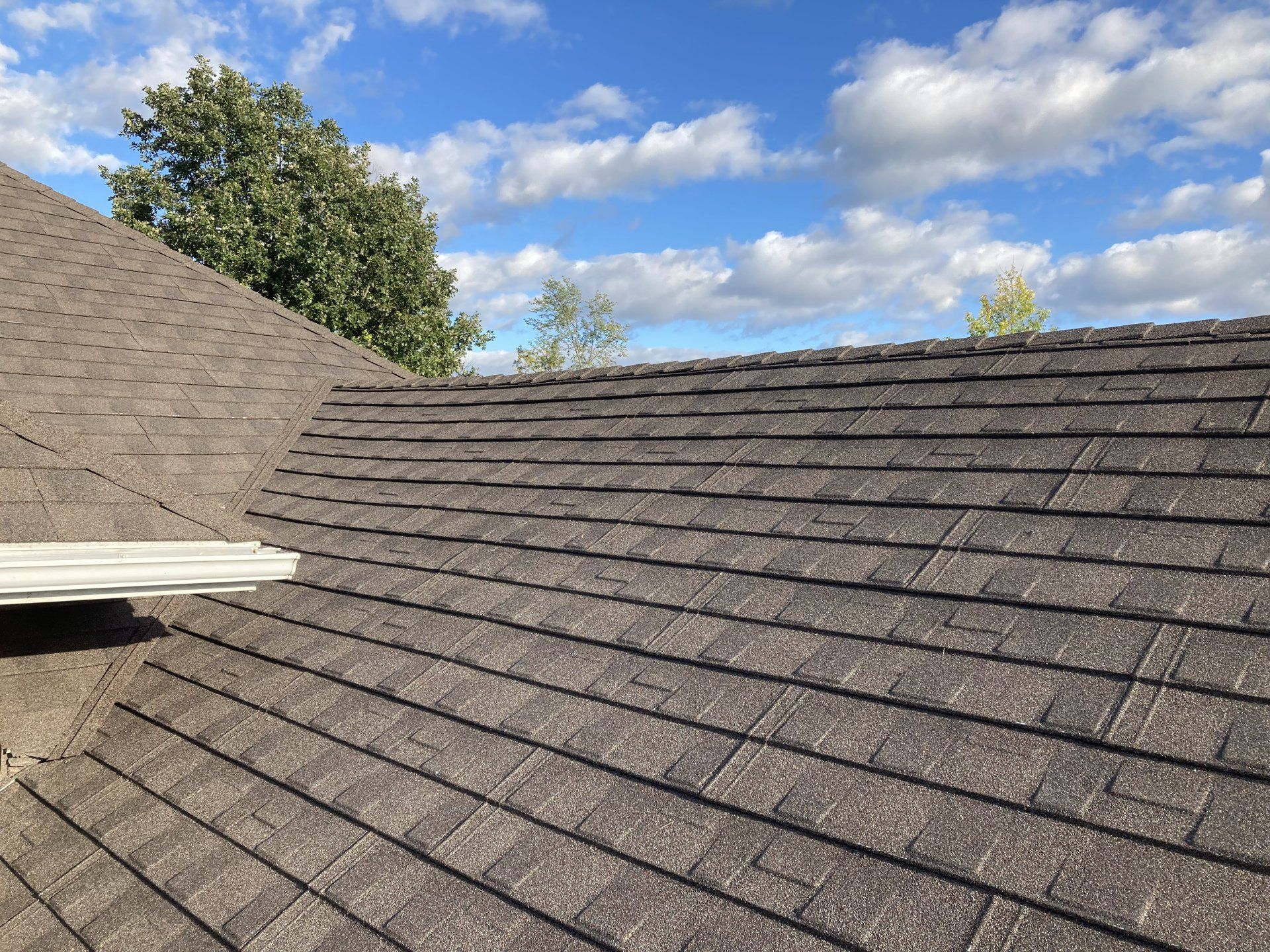 Shingled roof under blue sky with clouds and trees in the background