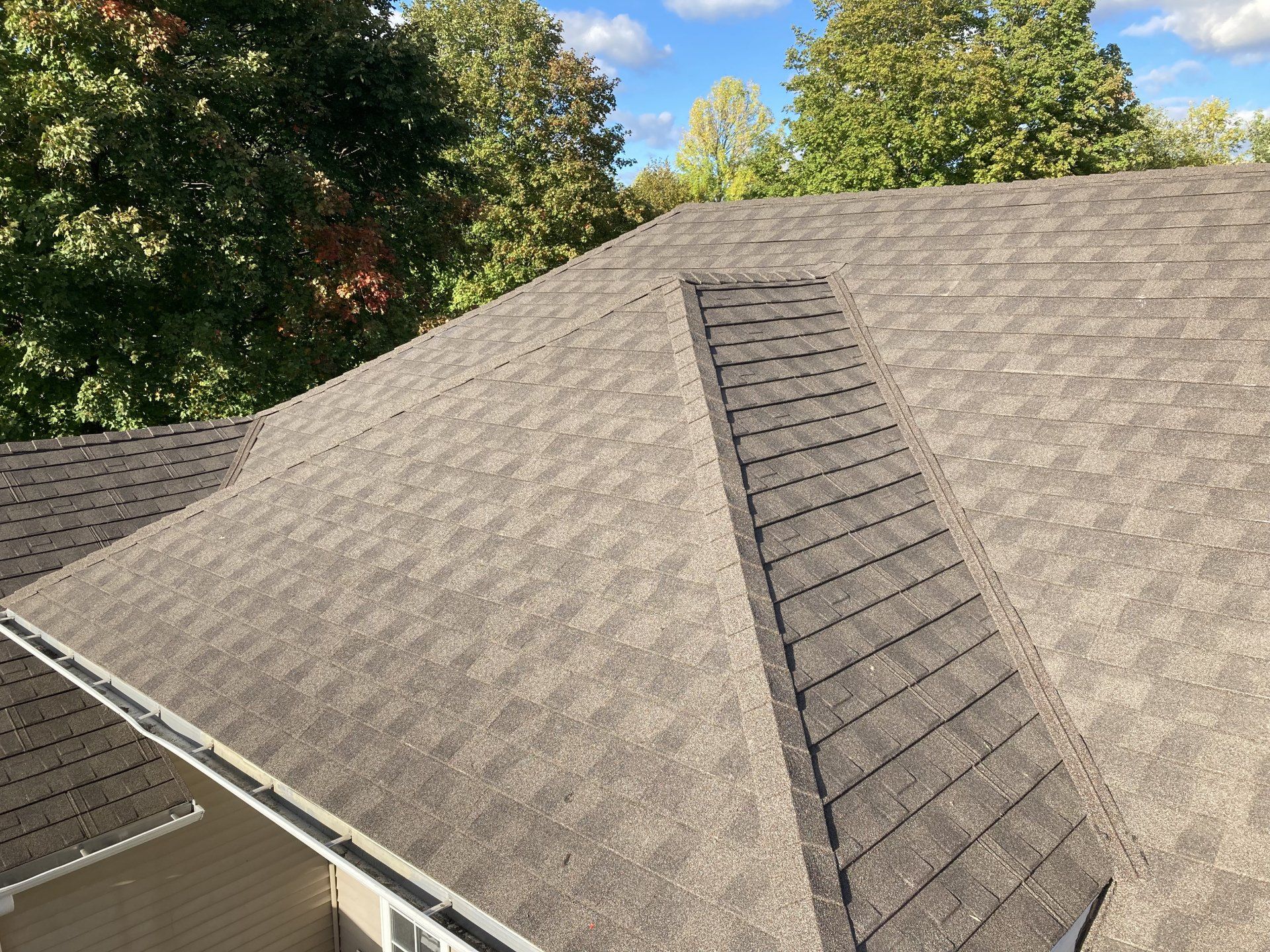 Roof with gray shingles and a dark ridge vent, surrounded by green trees under a blue sky
