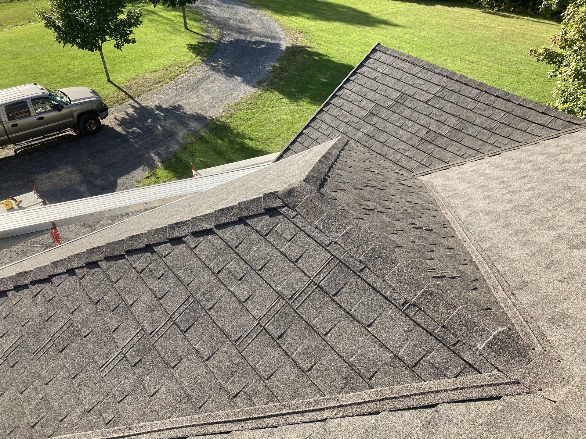 Aerial view of a shingled roof with a chimney, driveway, parked truck, and green yard below.