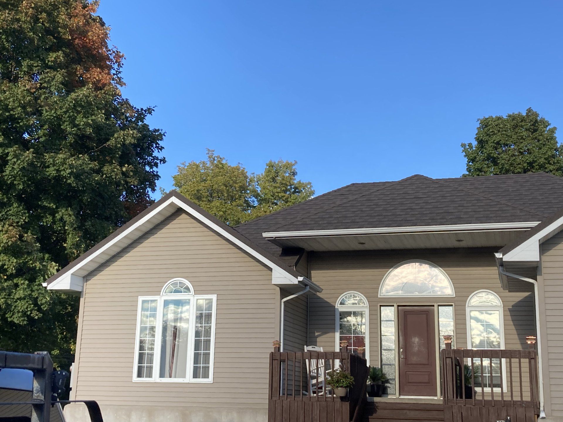 Suburban beige house with a brown roof, white trim, front porch, and trees under a blue sky