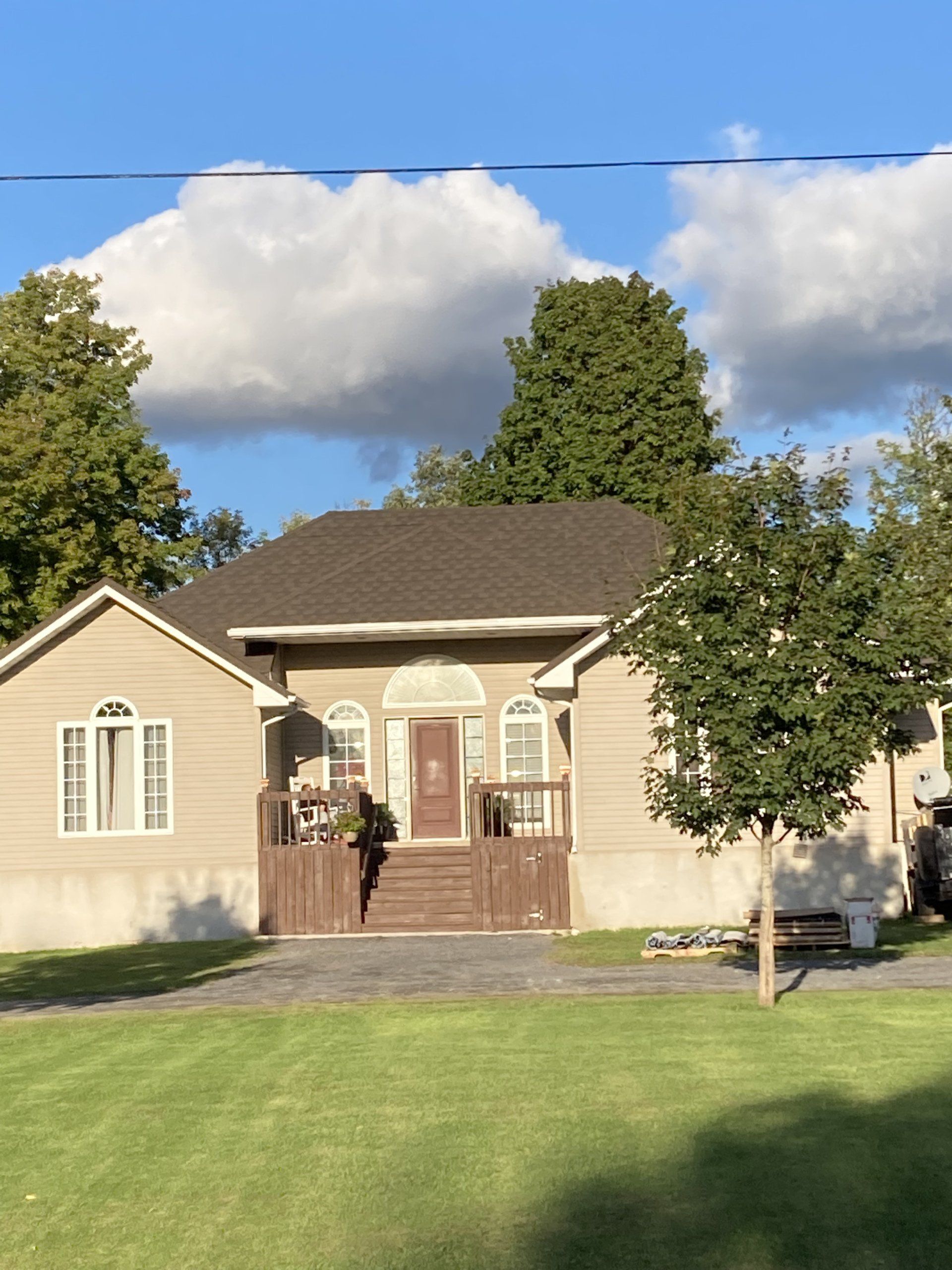 Single-story beige house with brown roof, front porch, and a person standing at the front door under a blue sky