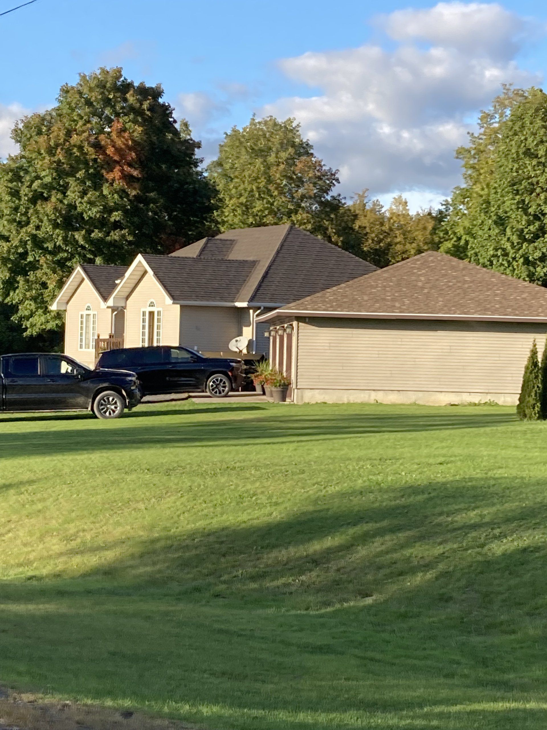 Suburban house with a large green lawn, trees, and two parked vehicles under a blue sky.