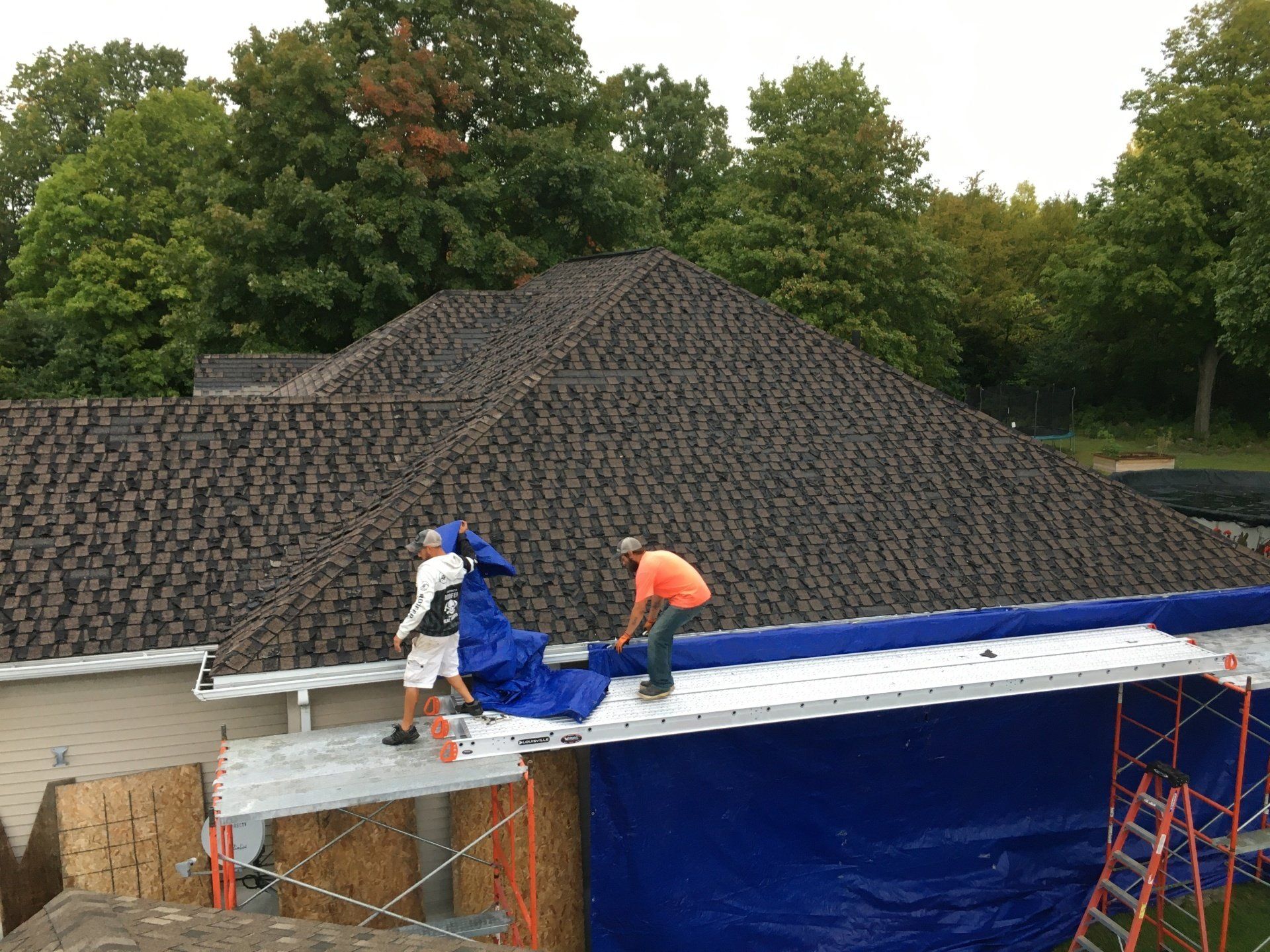 Workers on a roof under construction, installing blue tarps beneath a dark shingle roof.