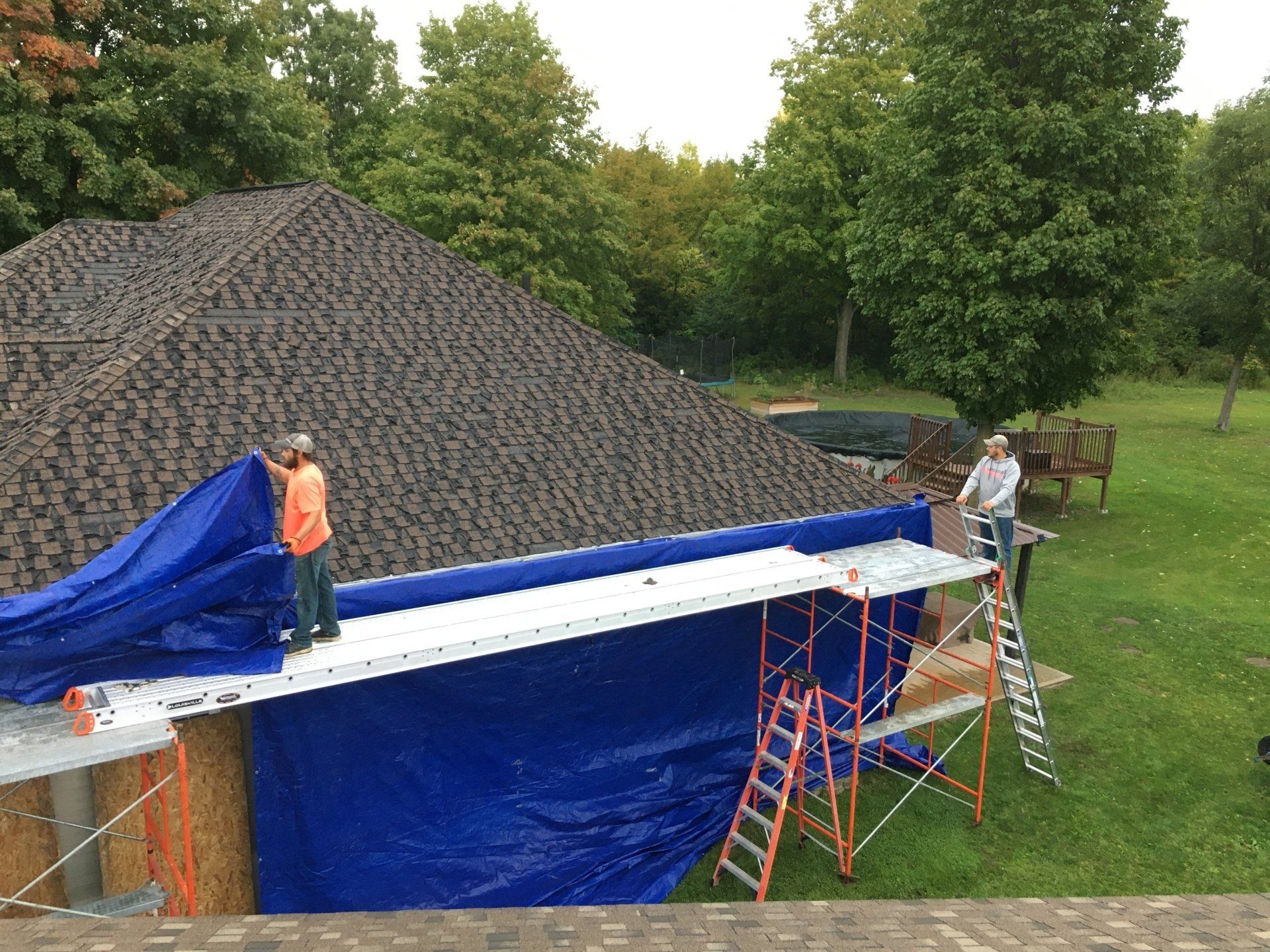 Workers on a house roof installing blue tarps beside a large pile of shingles