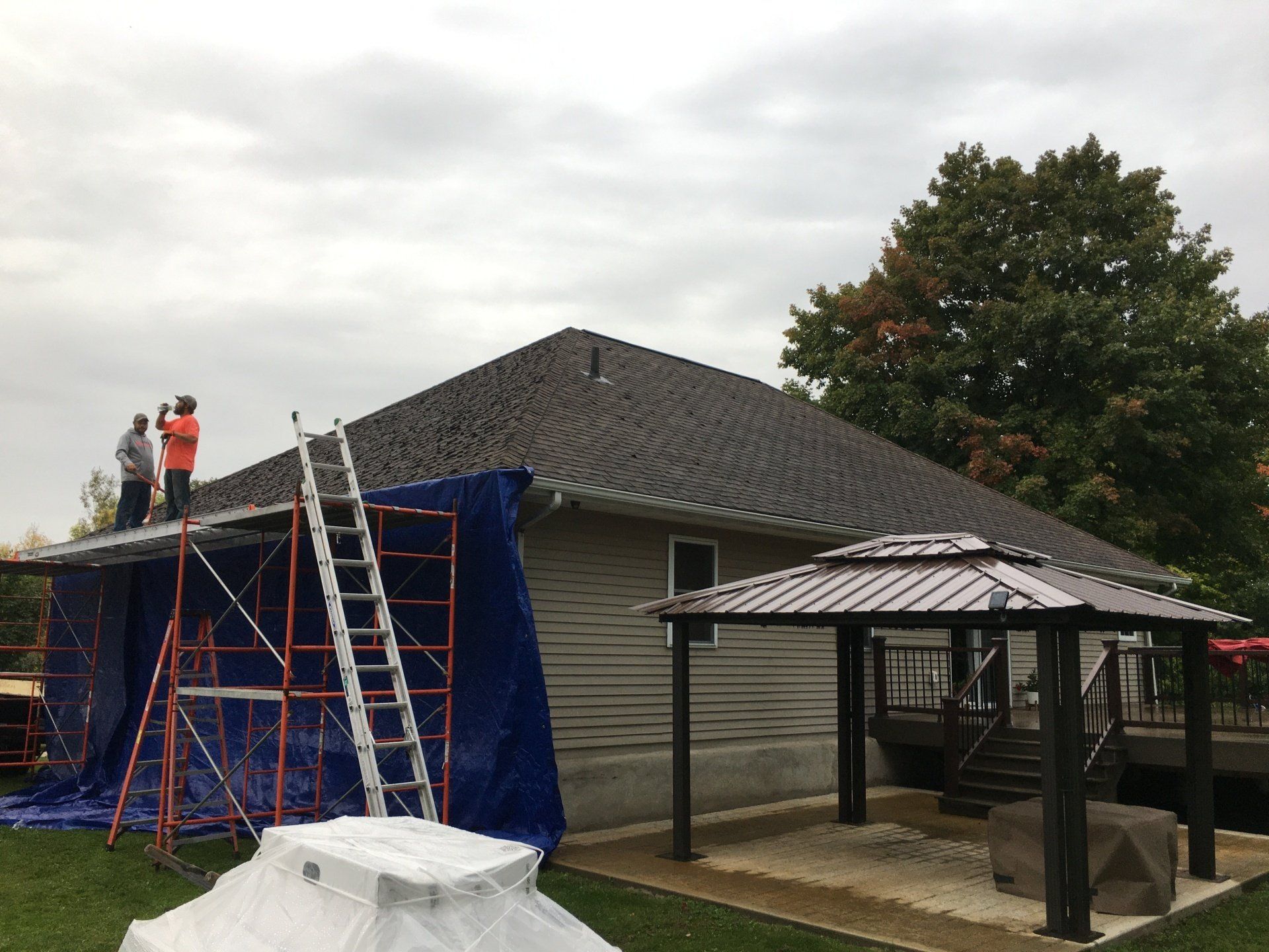Workers on a ladder reroofing a house beside a patio, with blue tarp and overcast sky.