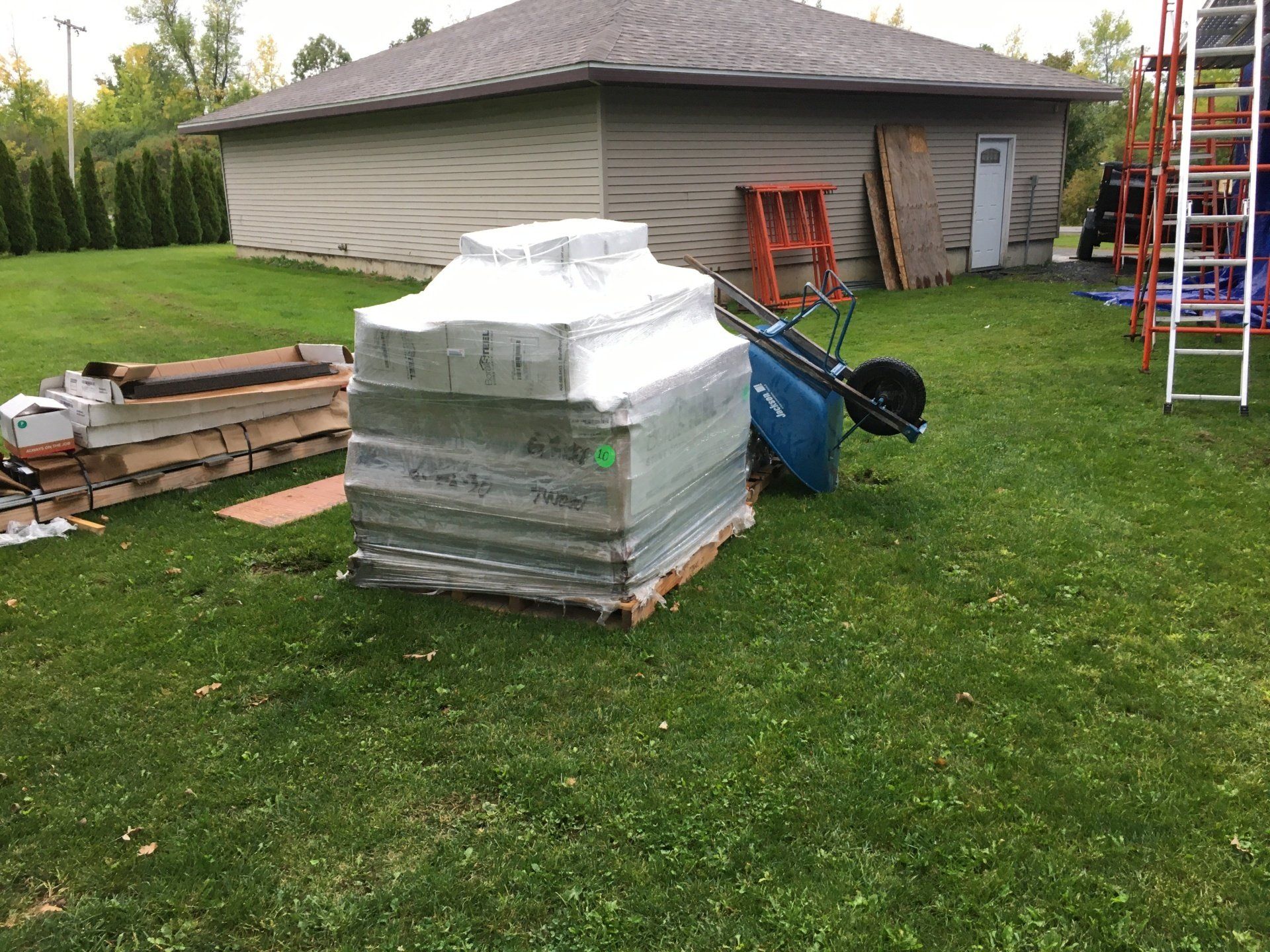 Backyard with stacked building materials, wheelbarrow, ladder, and tools beside a house under construction