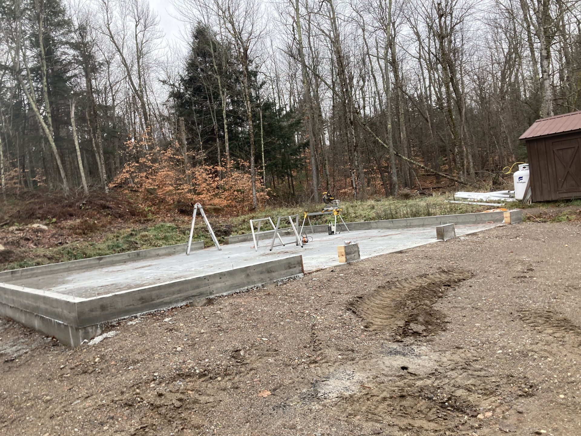 Concrete foundation slab in a wooded yard with muddy ground and a small shed nearby