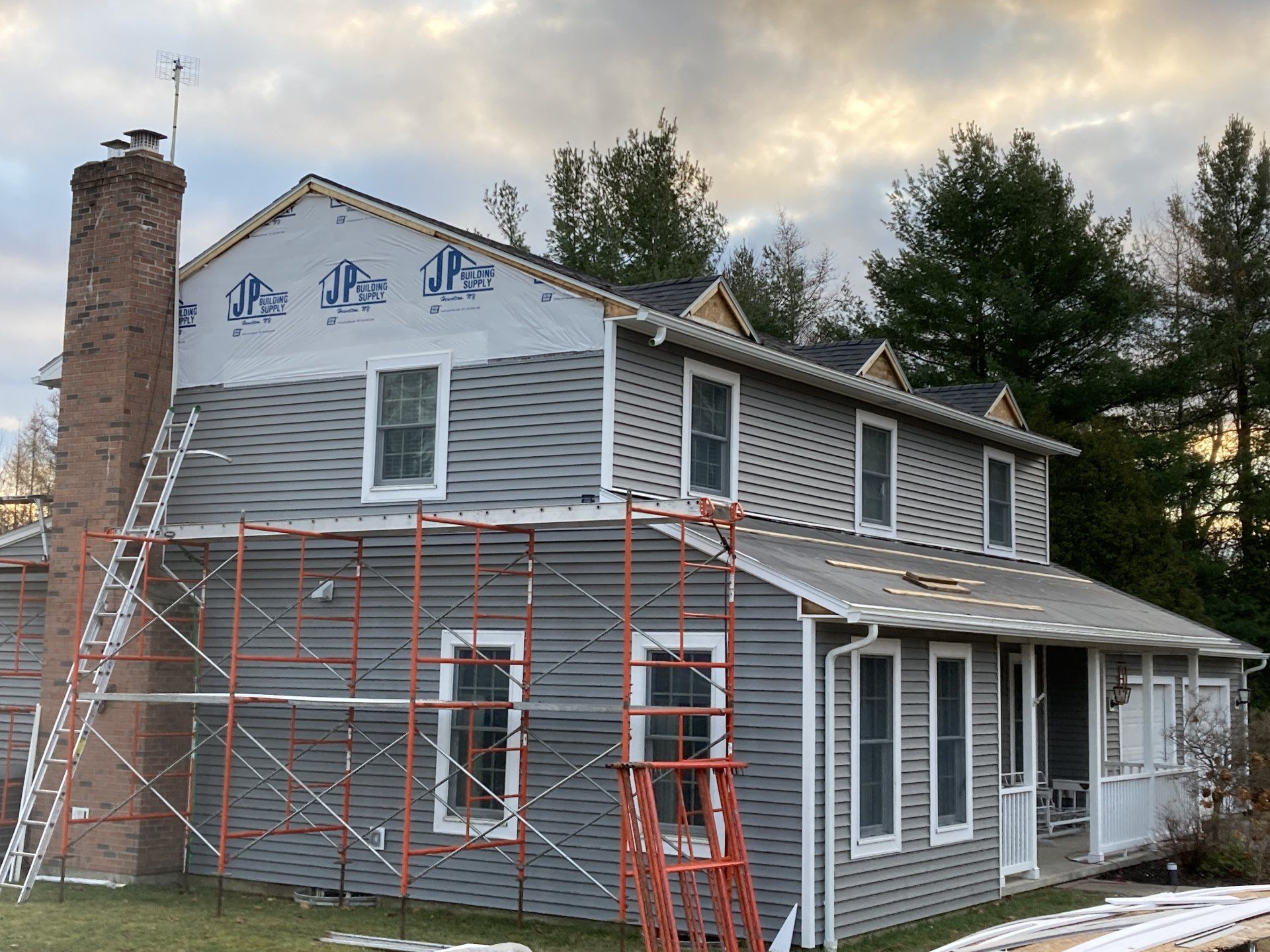 Two-story house under renovation with scaffolding, gray siding, white trim, and a brick chimney at sunset