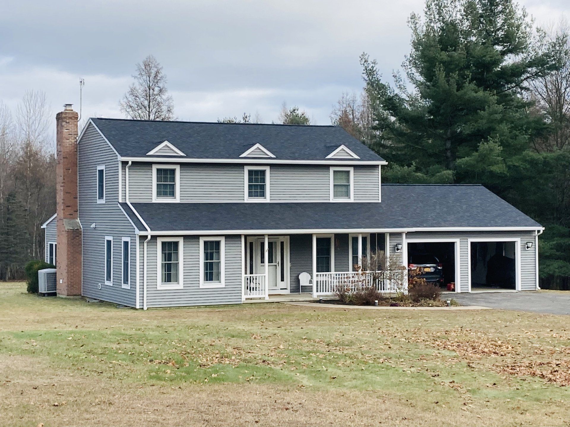 Gray two-story house with white trim, front porch, attached garage, and large lawn in a wooded area