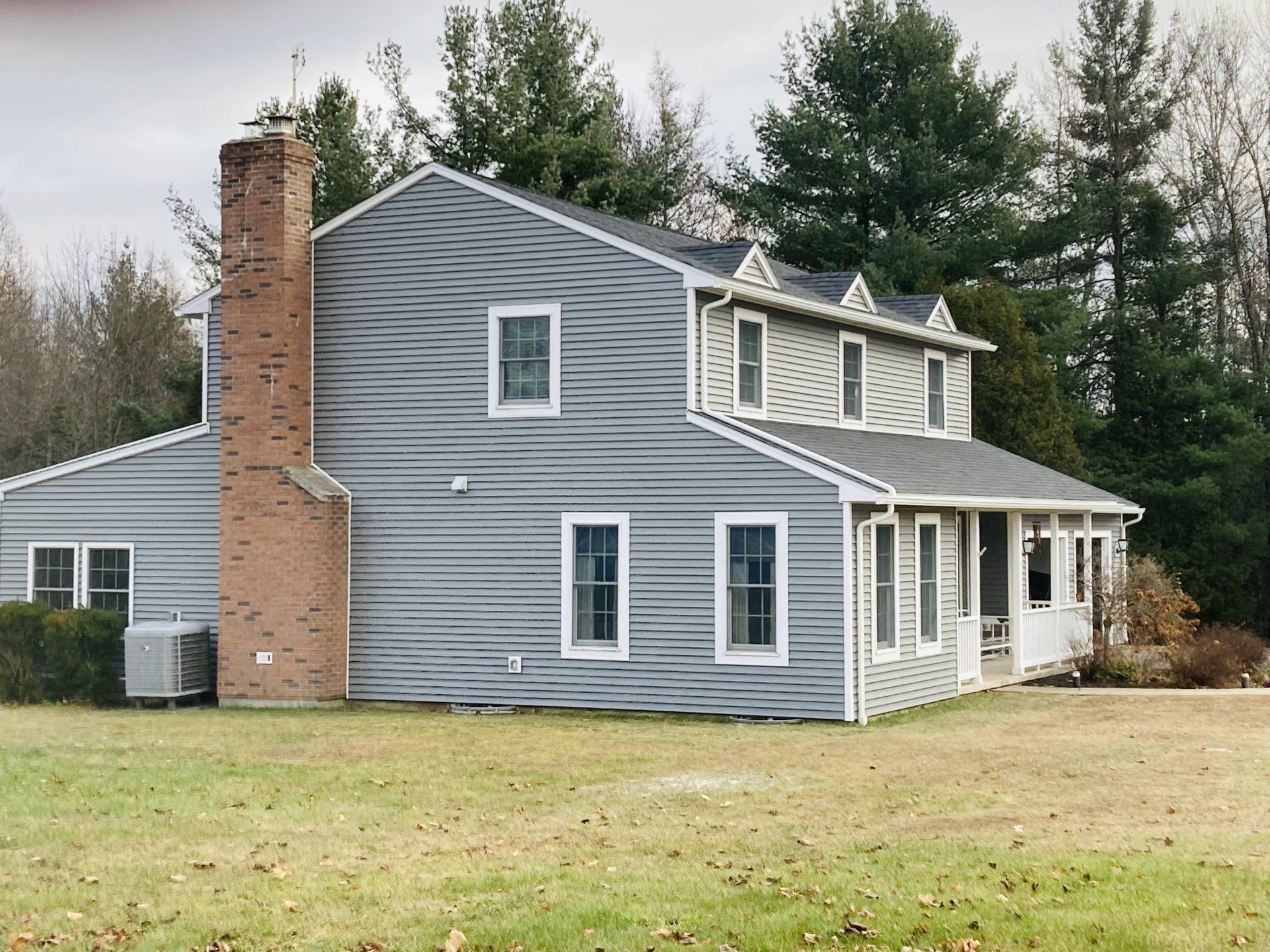 Gray two-story house with white trim, brick chimney, and covered porch on a grassy lot surrounded by trees