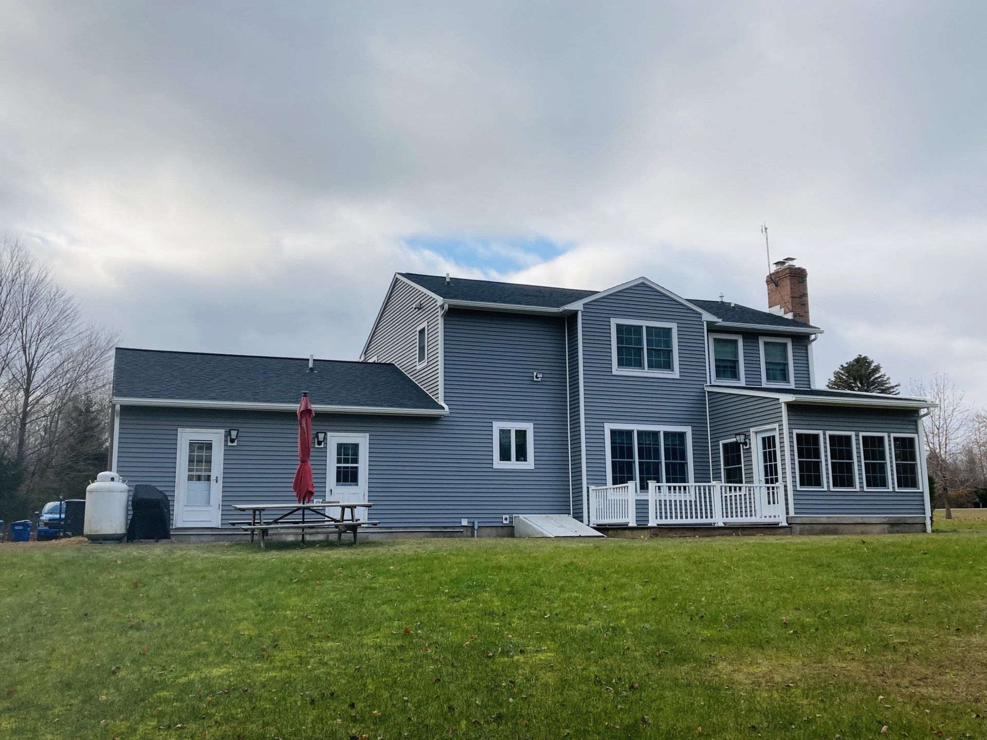Gray house with white trim and a porch, set on a grassy lawn under an overcast sky