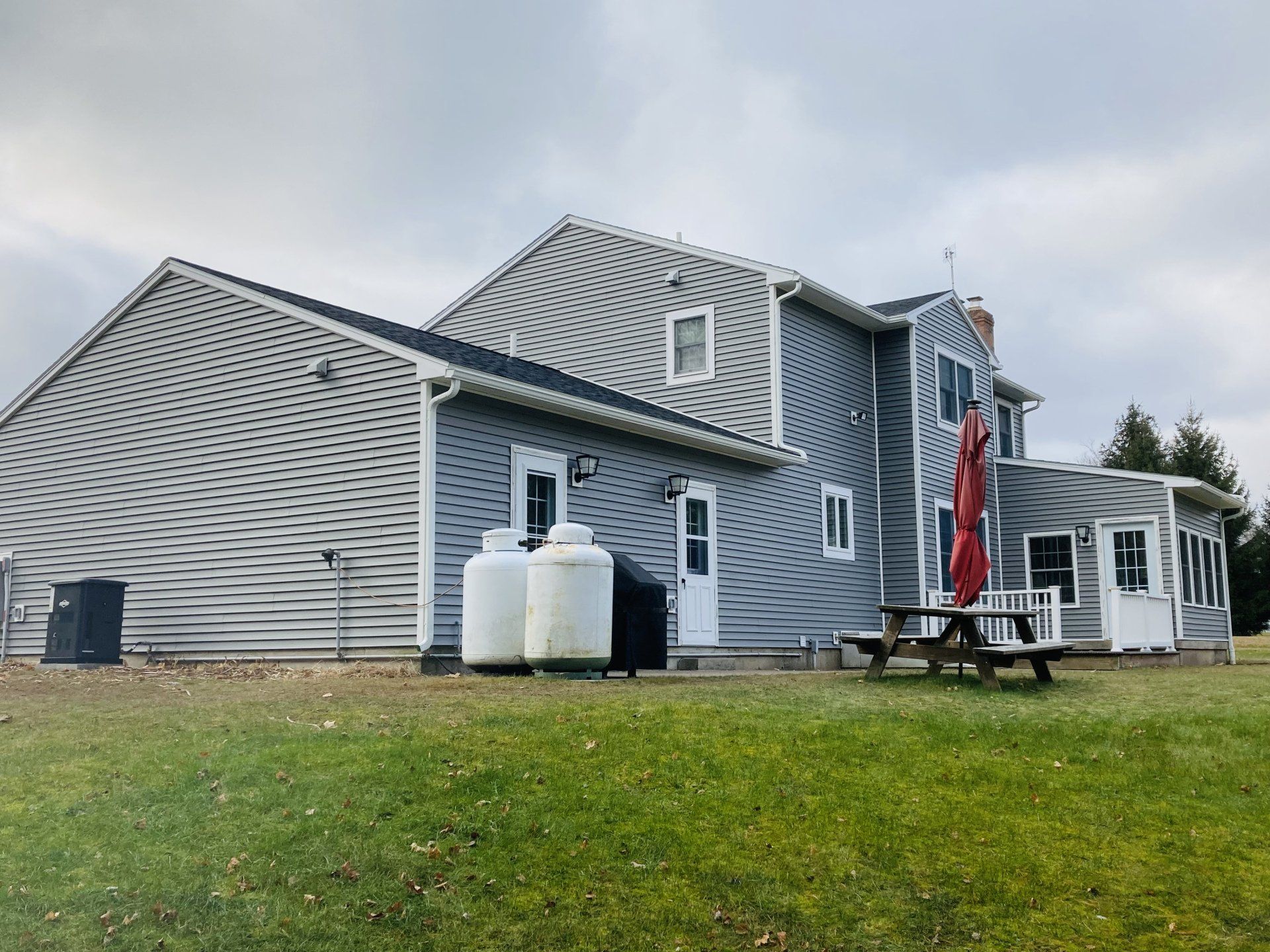 Gray house exterior with white trim, propane tanks, and a red patio umbrella on a grassy lawn under cloudy sky