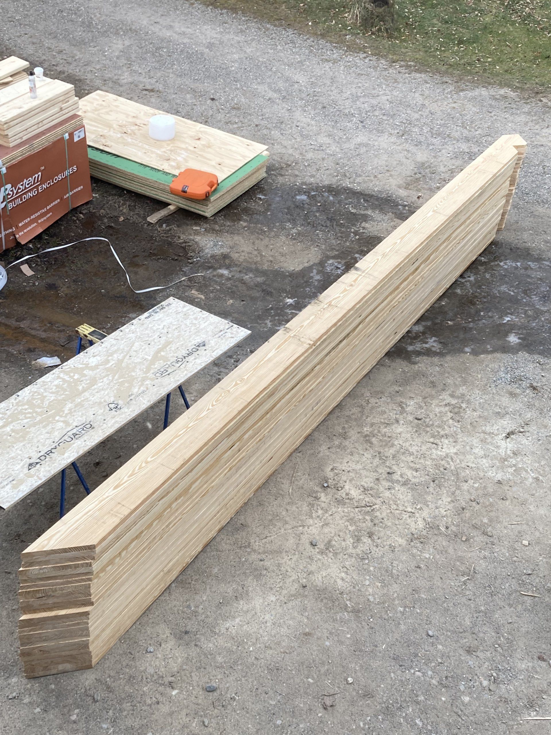 Long wooden board lying on a gravel surface beside stacked building materials.