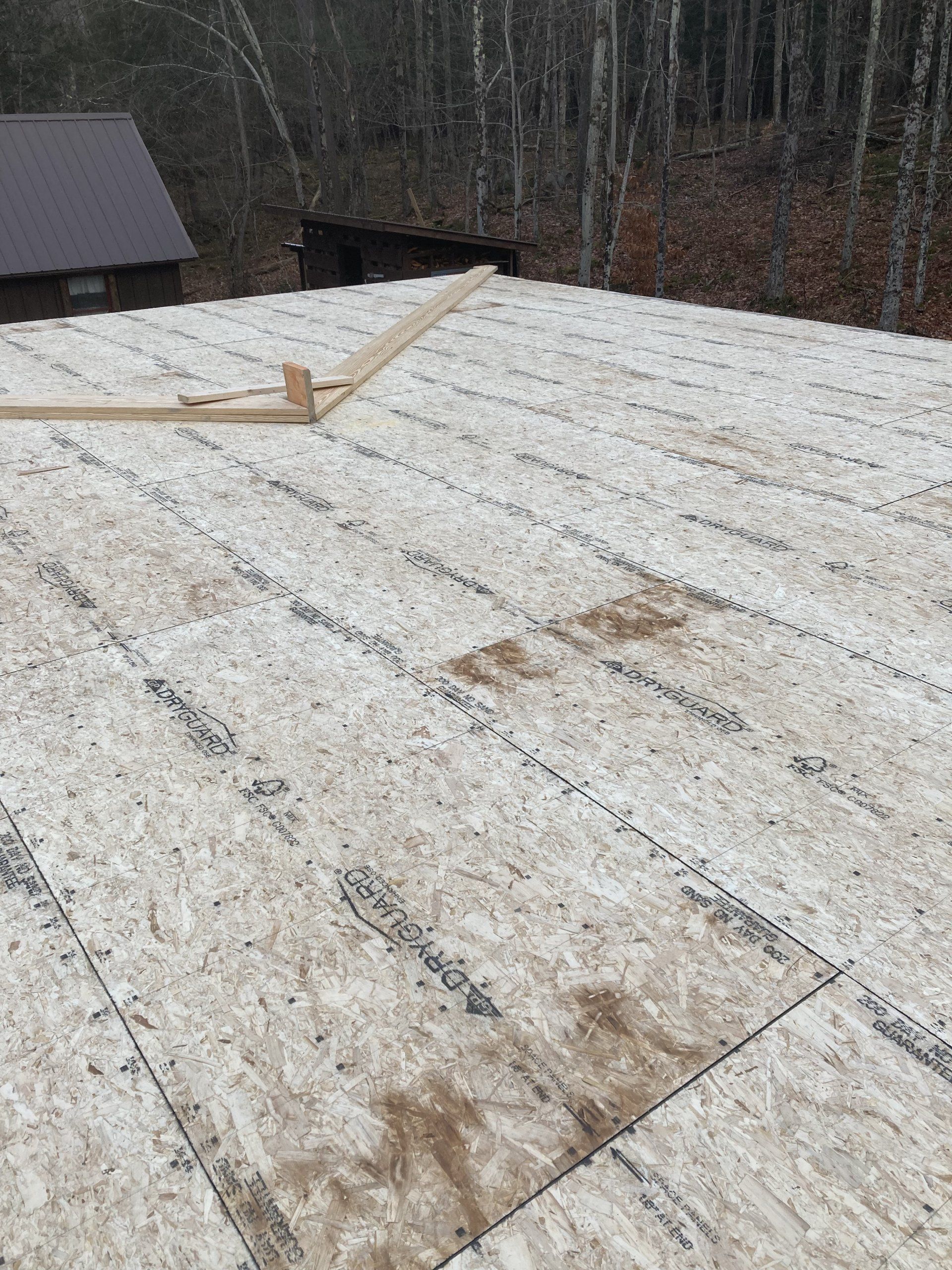 Snow-dusted roof under construction beside a wooded area, with a small cabin visible in the background