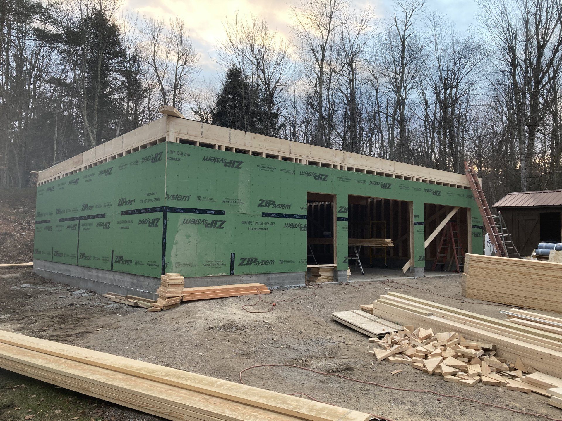 Partially built green house framed in a wooded lot with lumber and materials in front