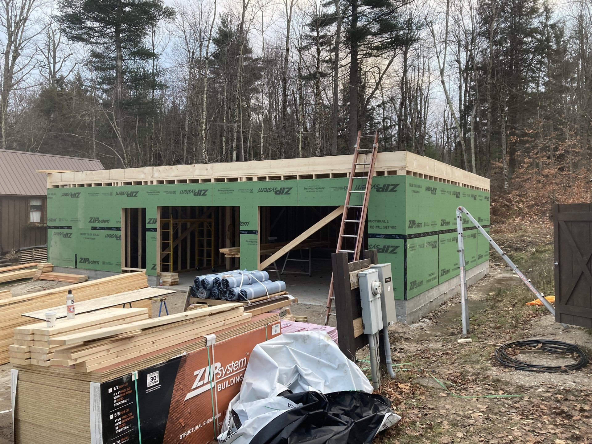 Partially built house with green sheathing, lumber stacks, and a ladder on a wooded lot