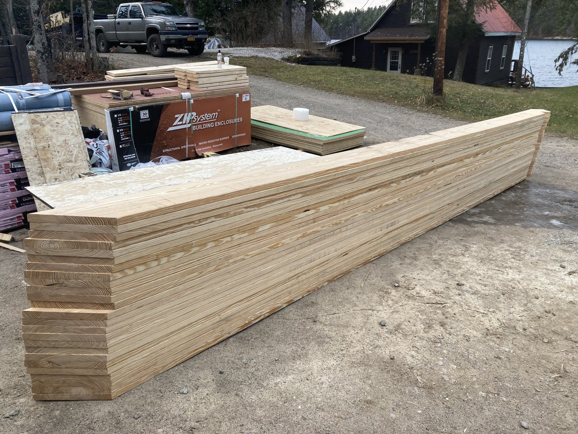 Stacks of long lumber boards piled outdoors beside a gravel driveway and parked truck.