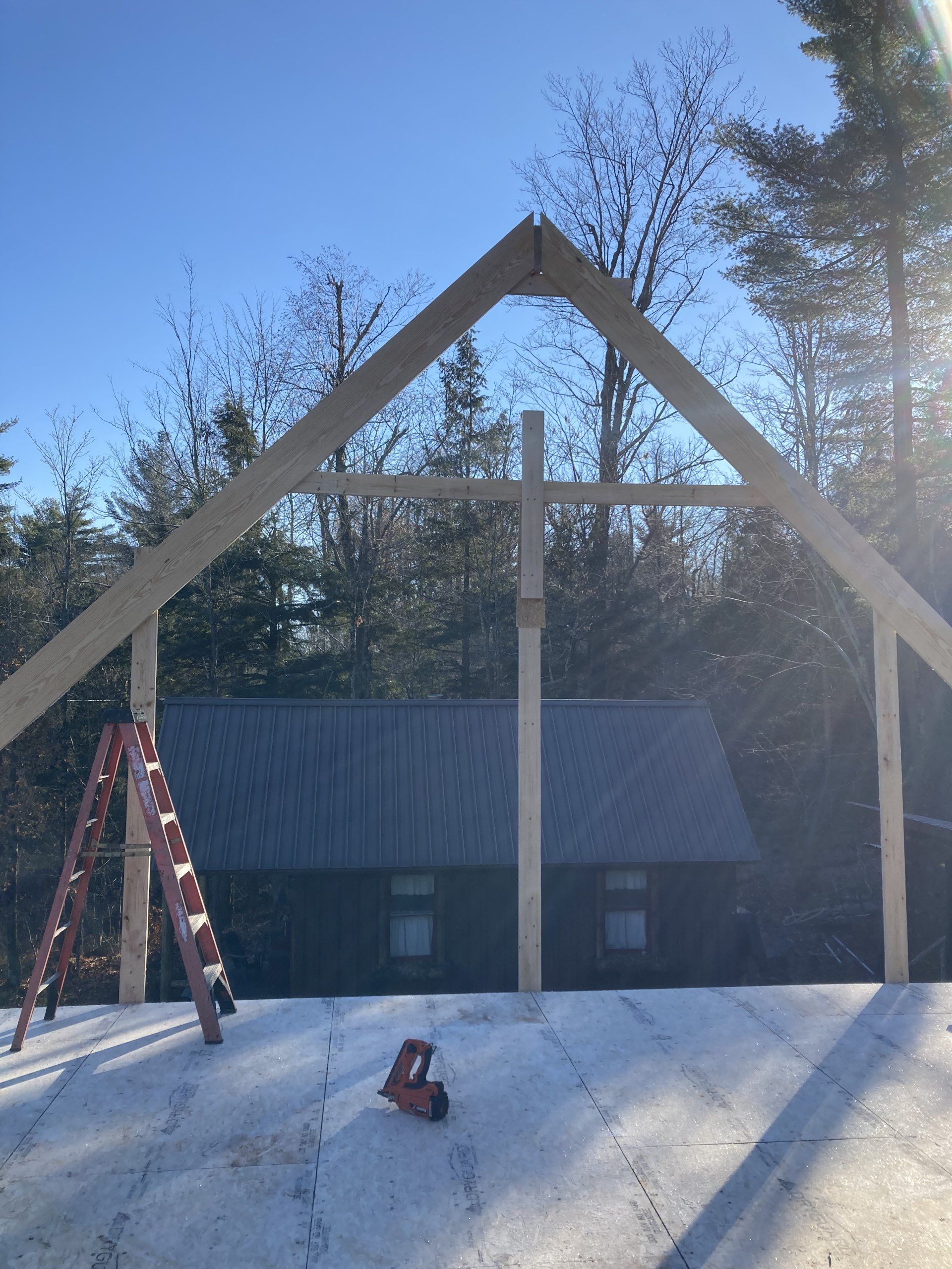 Large wooden A-frame under construction on a snowy site with a red ladder and a cabin in the background