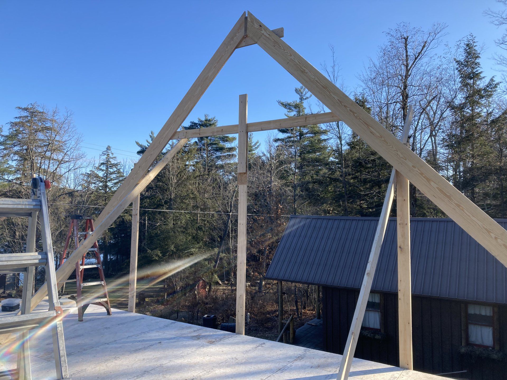 Wooden A-frame structure under construction beside a dark roofed cabin in a wooded area