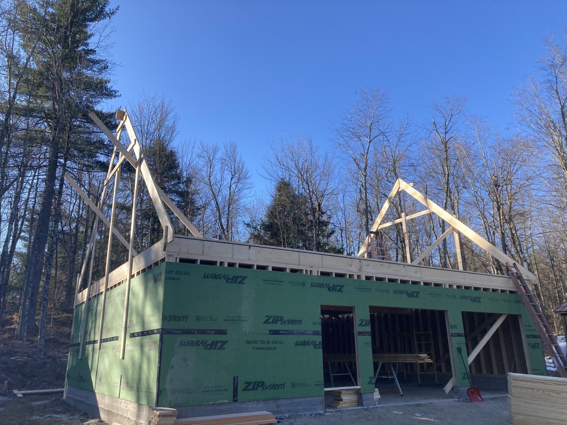Partially built house with green siding and roof trusses in a wooded area under a blue sky