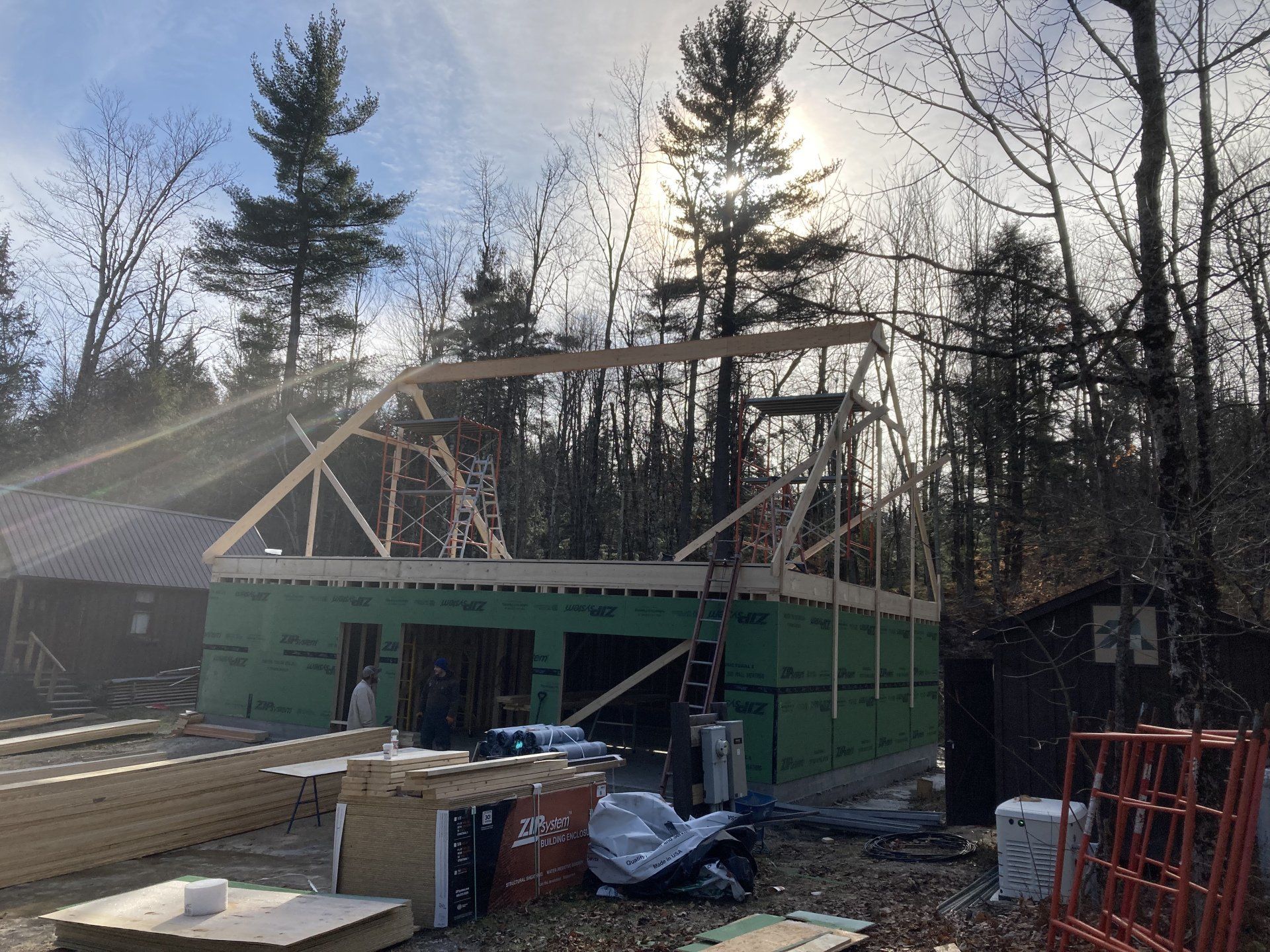 Partially built house with green walls and exposed roof framing in a wooded area, construction materials scattered nearby