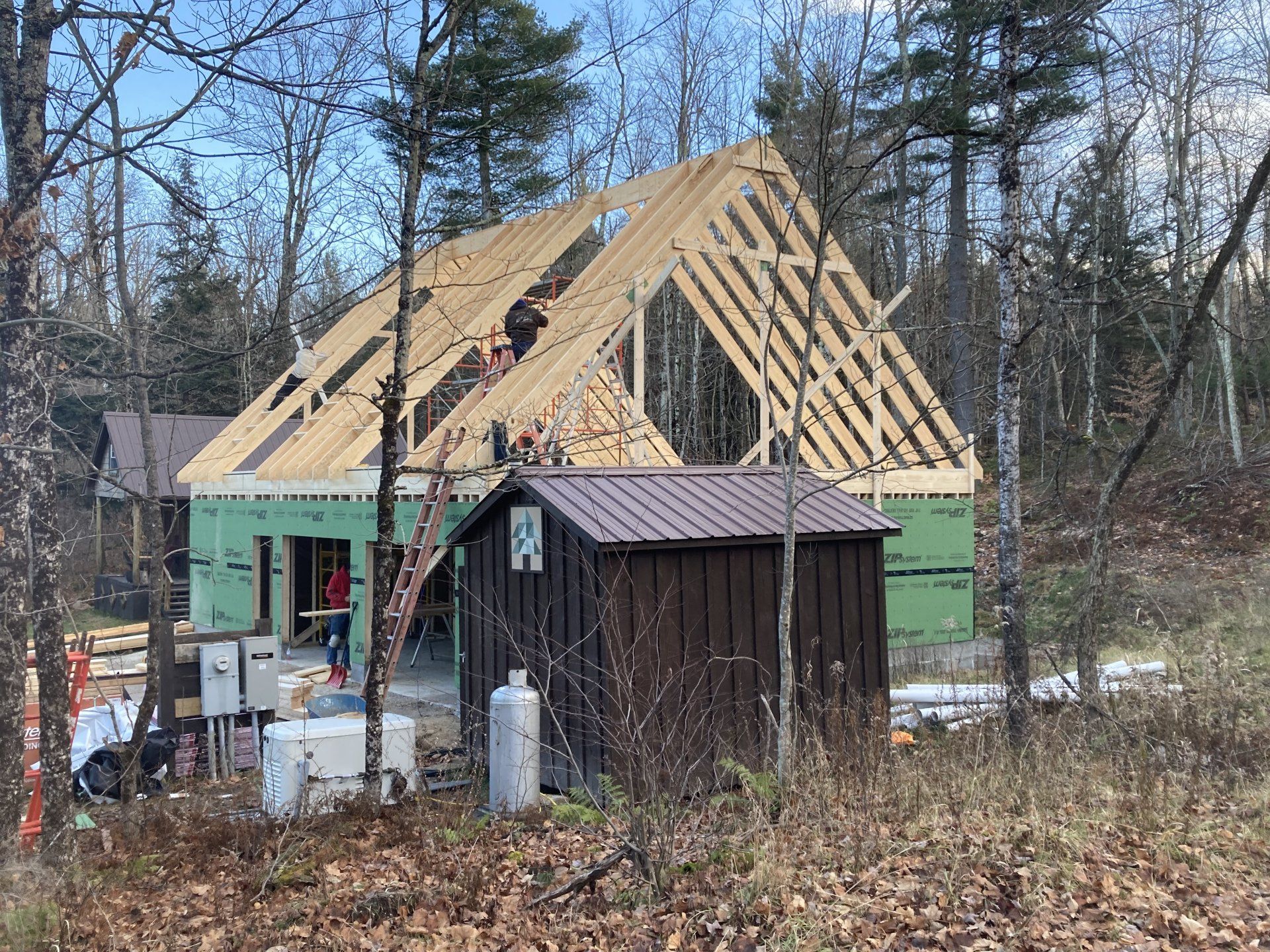 House under construction with exposed roof framing beside a shed in a wooded yard