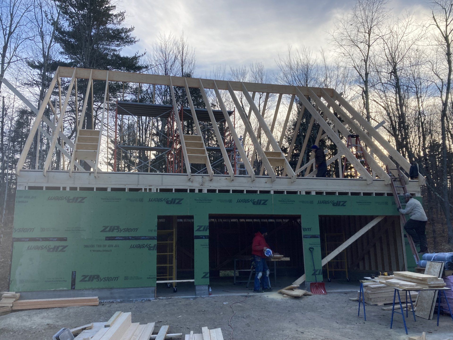 House under construction with roof framing, green sheathing, and workers on a ladder and inside.
