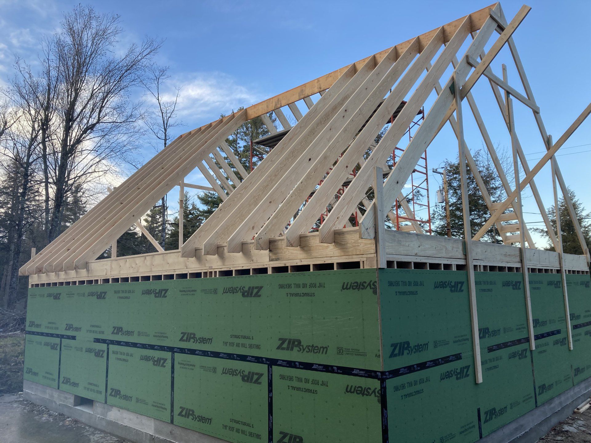 House under construction with roof trusses, wrapped in green insulation under a blue sky
