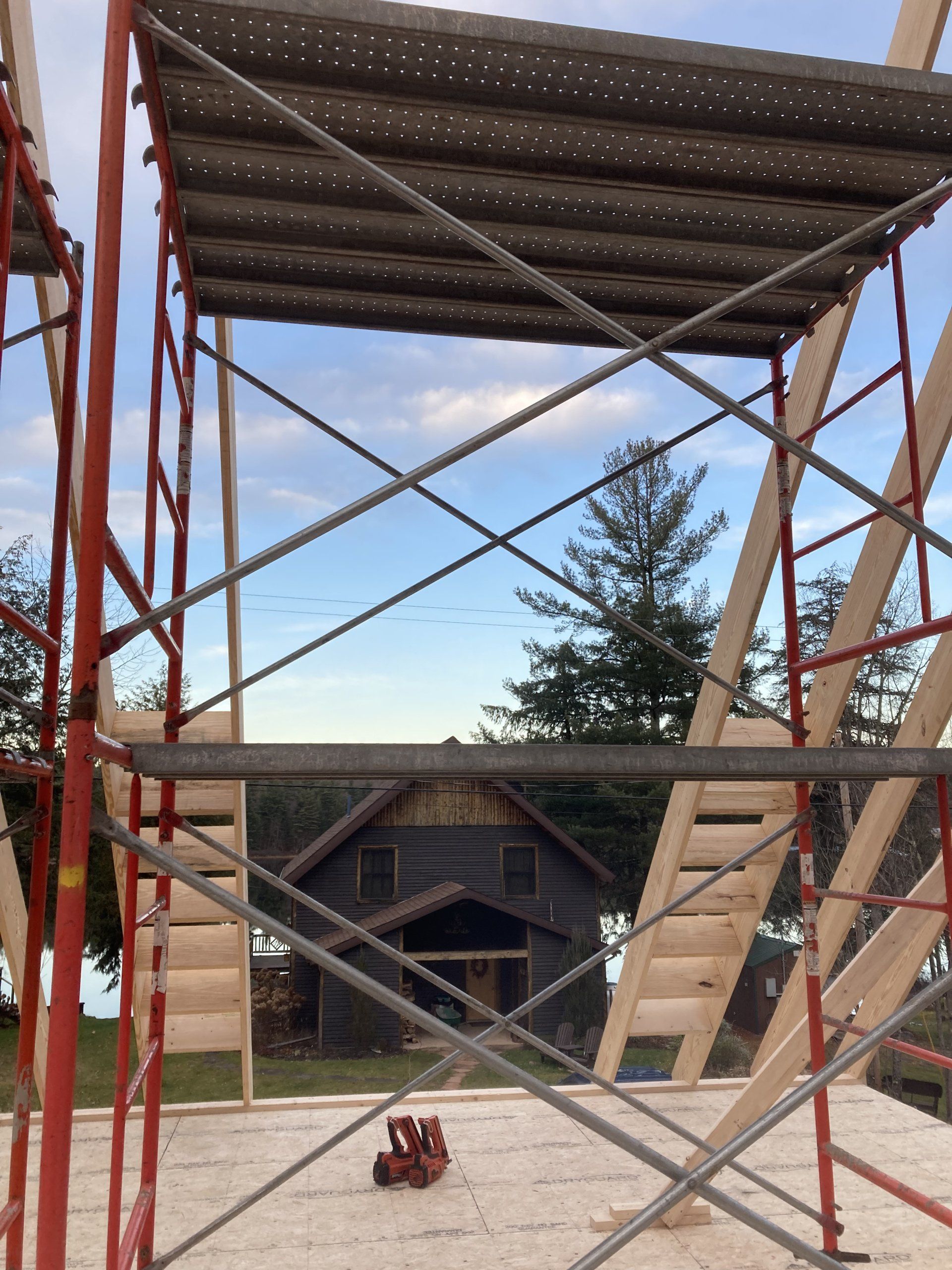 Red scaffolding tower with metal cross-bracing, viewed toward a small cabin and trees in the background.