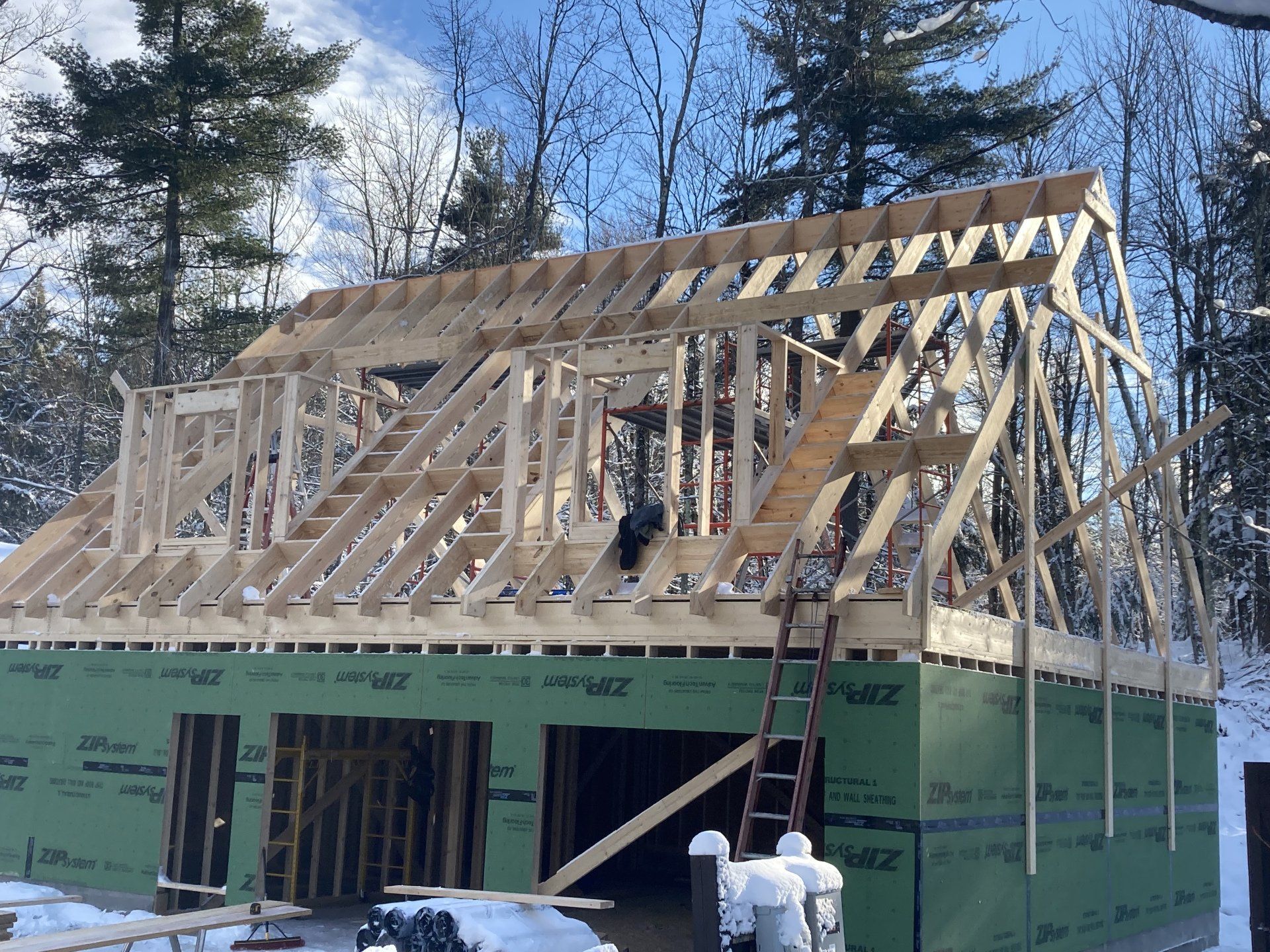 House under construction with exposed wooden framing and green sheathing, workers visible at the foundation