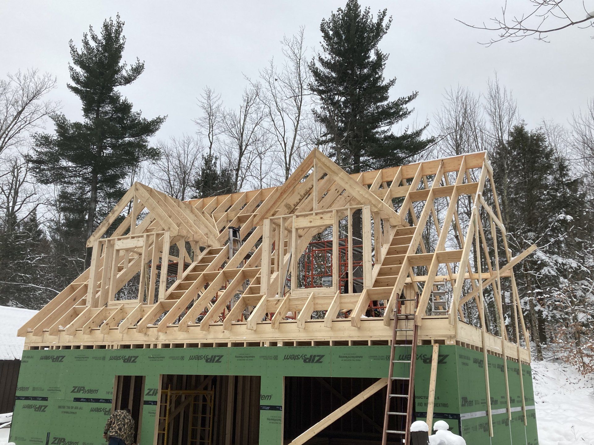Wood-framed house under construction on a snowy lot, with green sheathing and tall trees behind