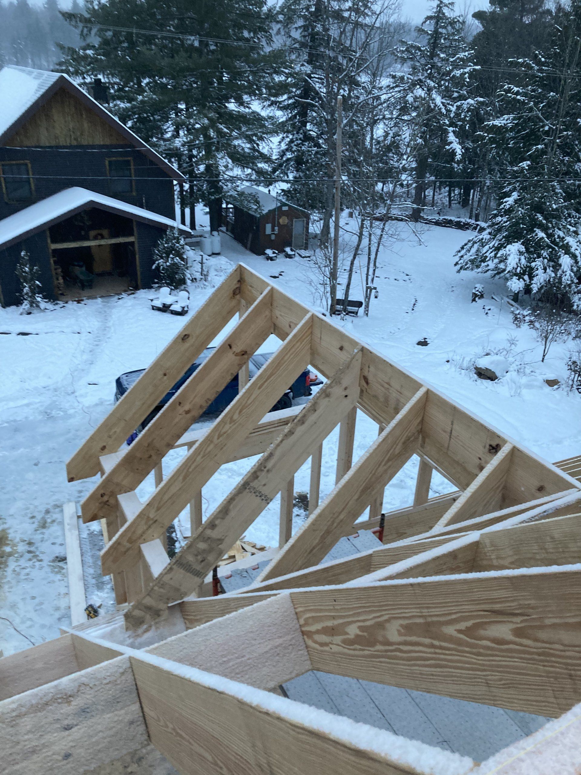Snowy yard with unfinished wooden roof framing beside a dark cabin and pine trees