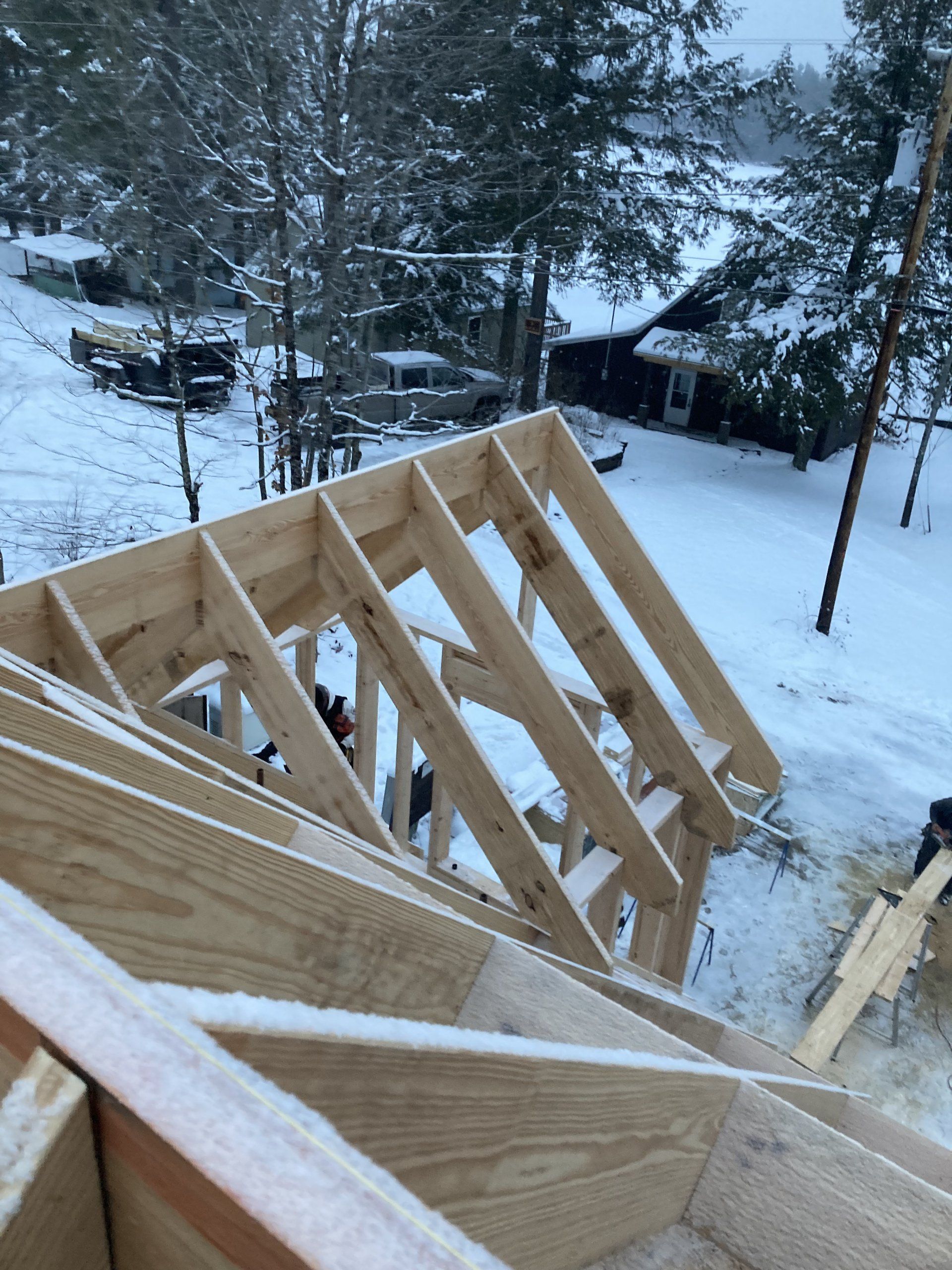 Snow-covered wooden roof framing on a house with trees and a cabin in the background