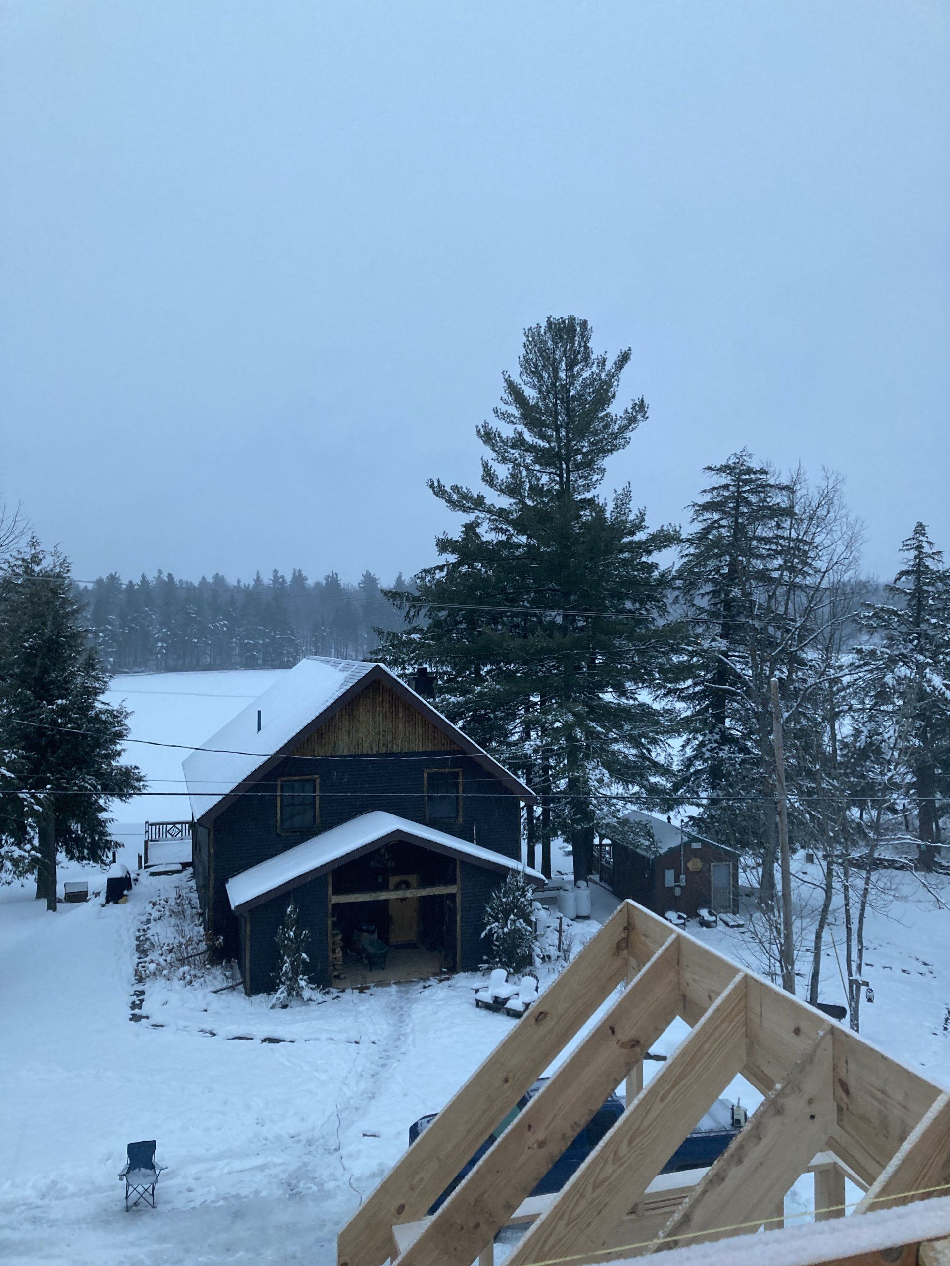 Snowy rural cabin with wood-framed roof under construction, pine trees, and overcast winter sky